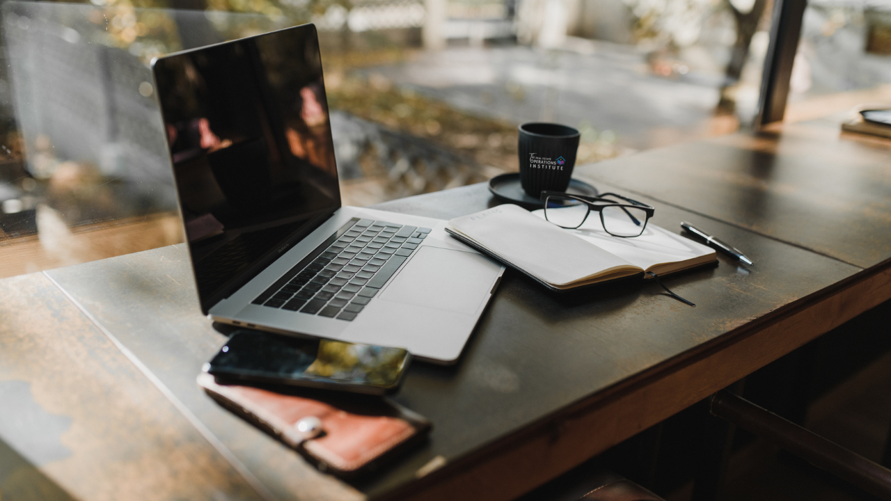 image of a laptop on a wooden table outside, along with an open notebook, 2 cellphones, eye glasses, and black mug.