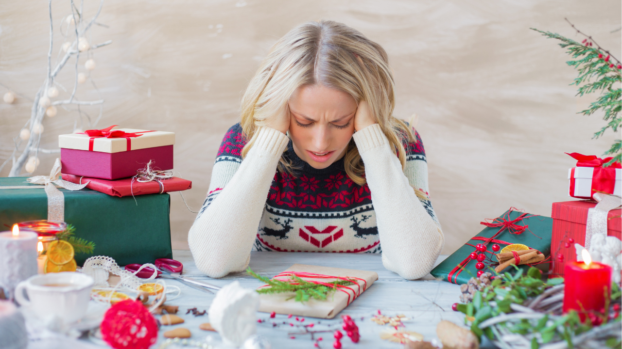 a woman sitting with her hands covering her ears, visually stressed out surrounded by all things christmas: presents, ornaments, cookies, coffee
