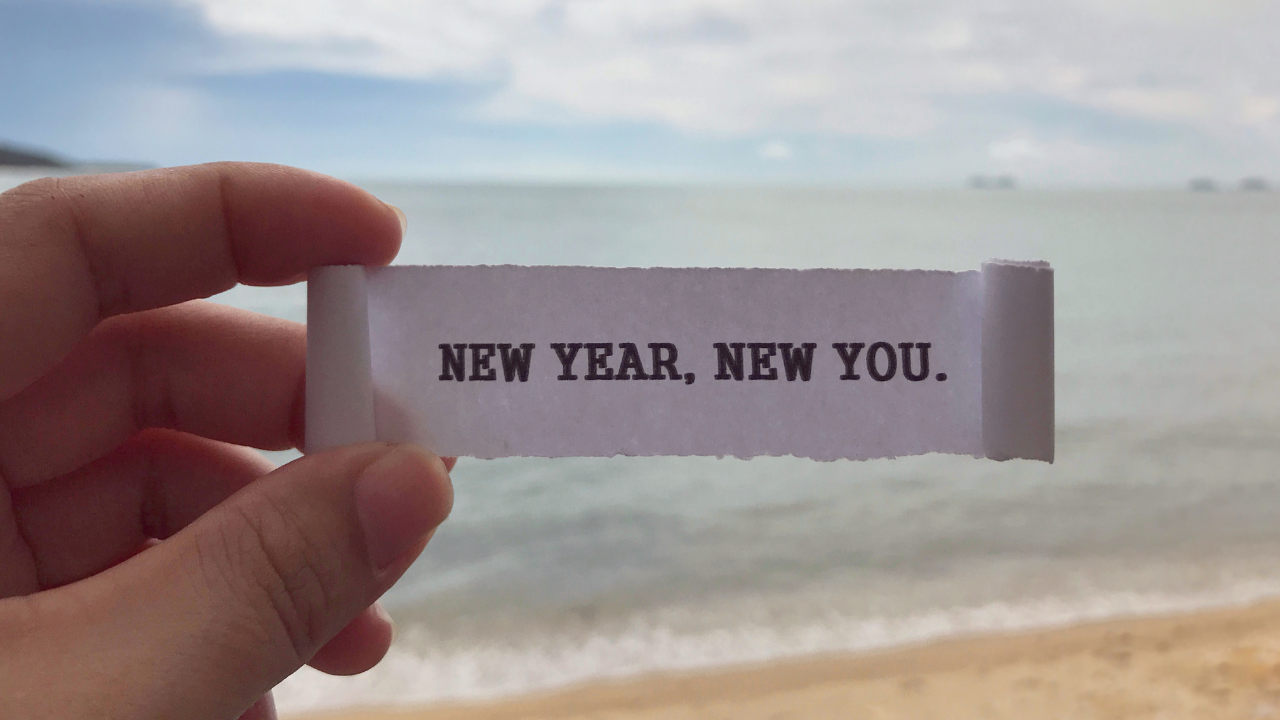 Ladies hand holding up a small note that reads "New Year, New You" on the beach