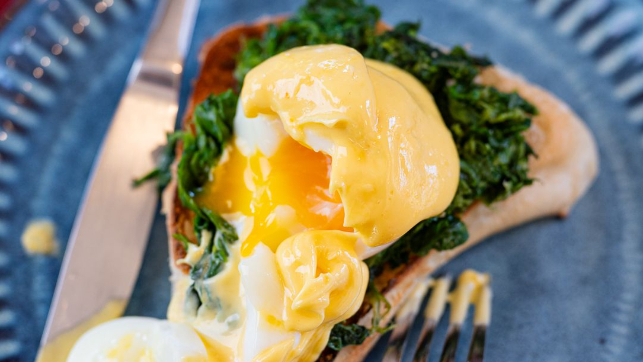 creamy hollandaise sauce being poured over eggs benedict with golden egg yolk visible