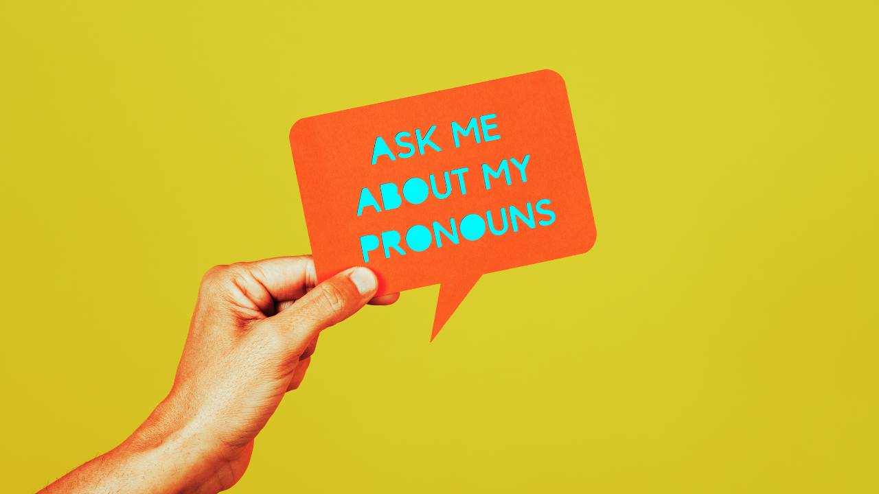 A hand holds up a sign that says "Ask me about my pronouns."