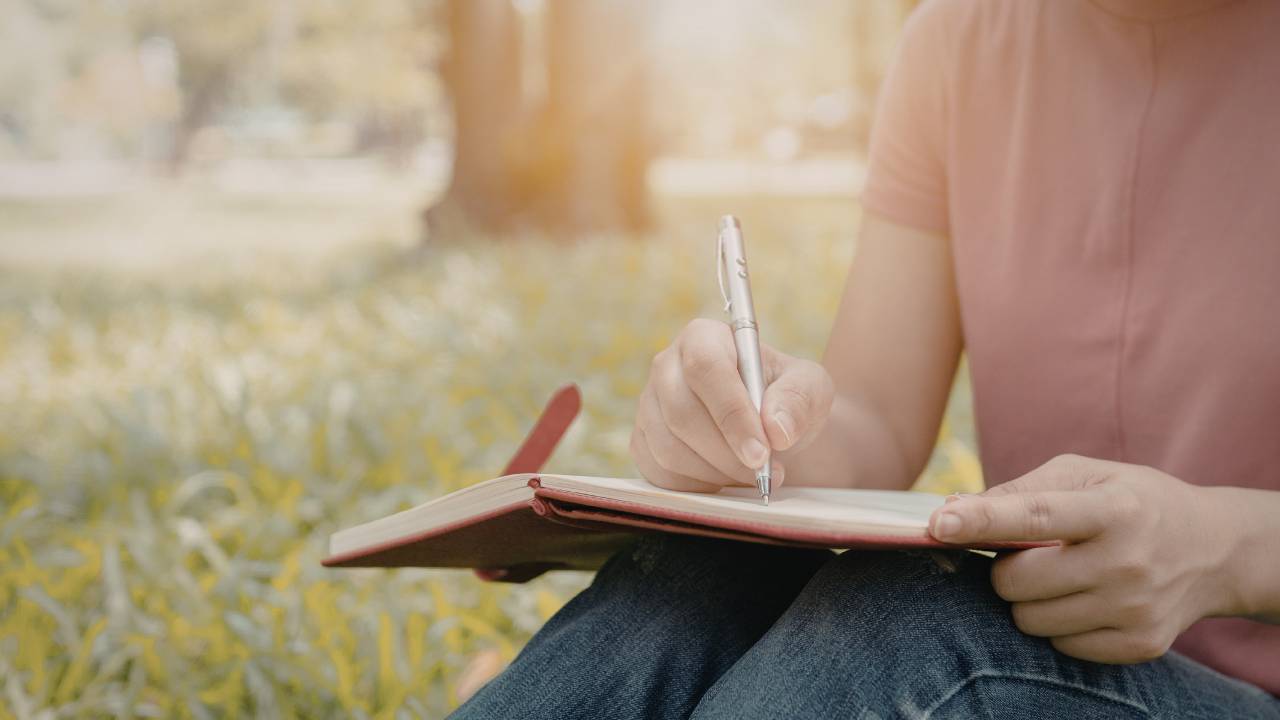 person sitting in a park with a pink shirt and jeans on, writing in a journal, grass and flowers in the park and a tree in distance, sun shining down