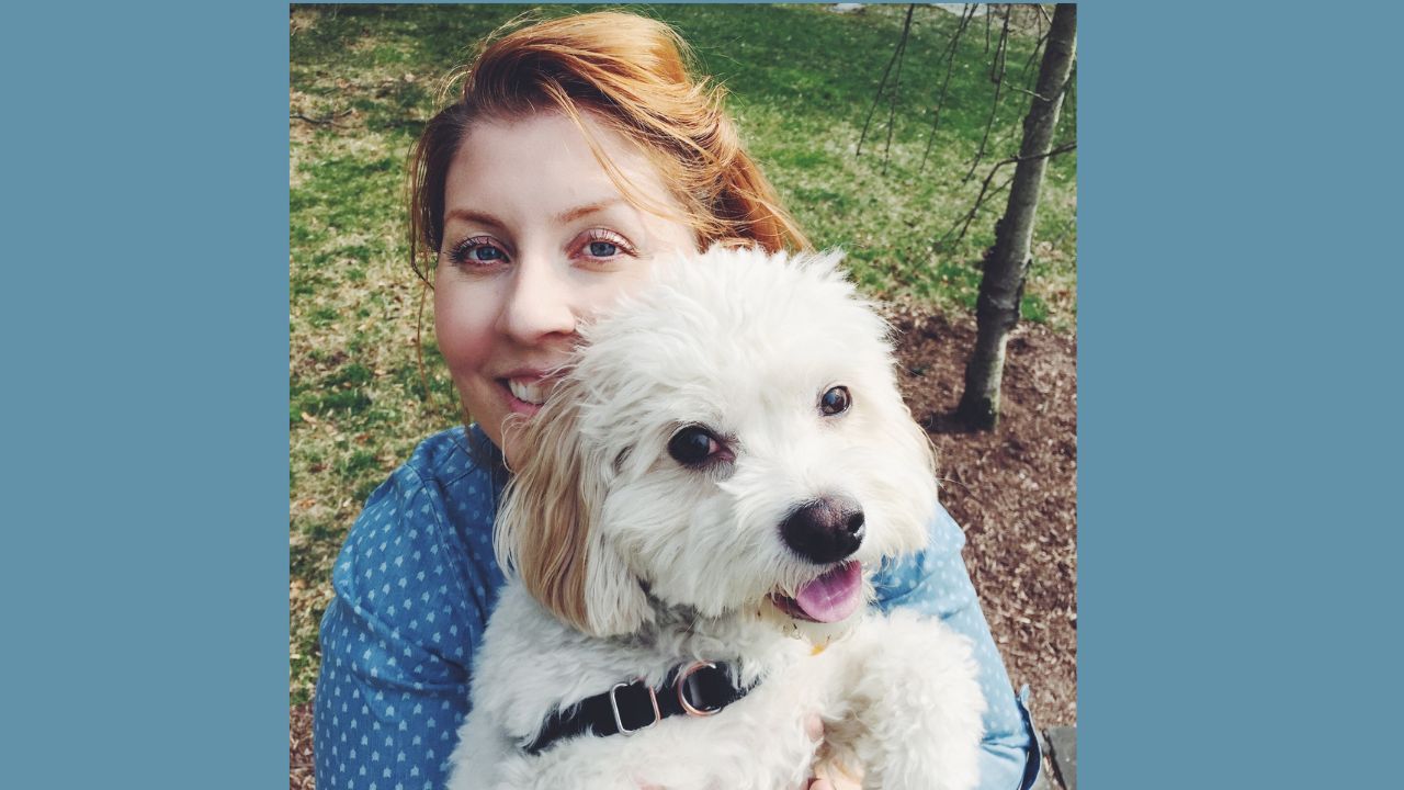 super cute white dog with dark eyes and a pink tongue being held by a smilling redheaded white woman