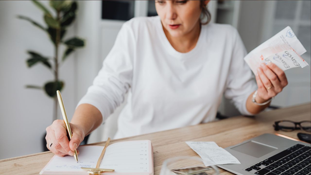 woman holding receipts in one hand and recording them in a journal with her other, wearing a white long sleeved shirt and sitting at a desk in front of her laptop