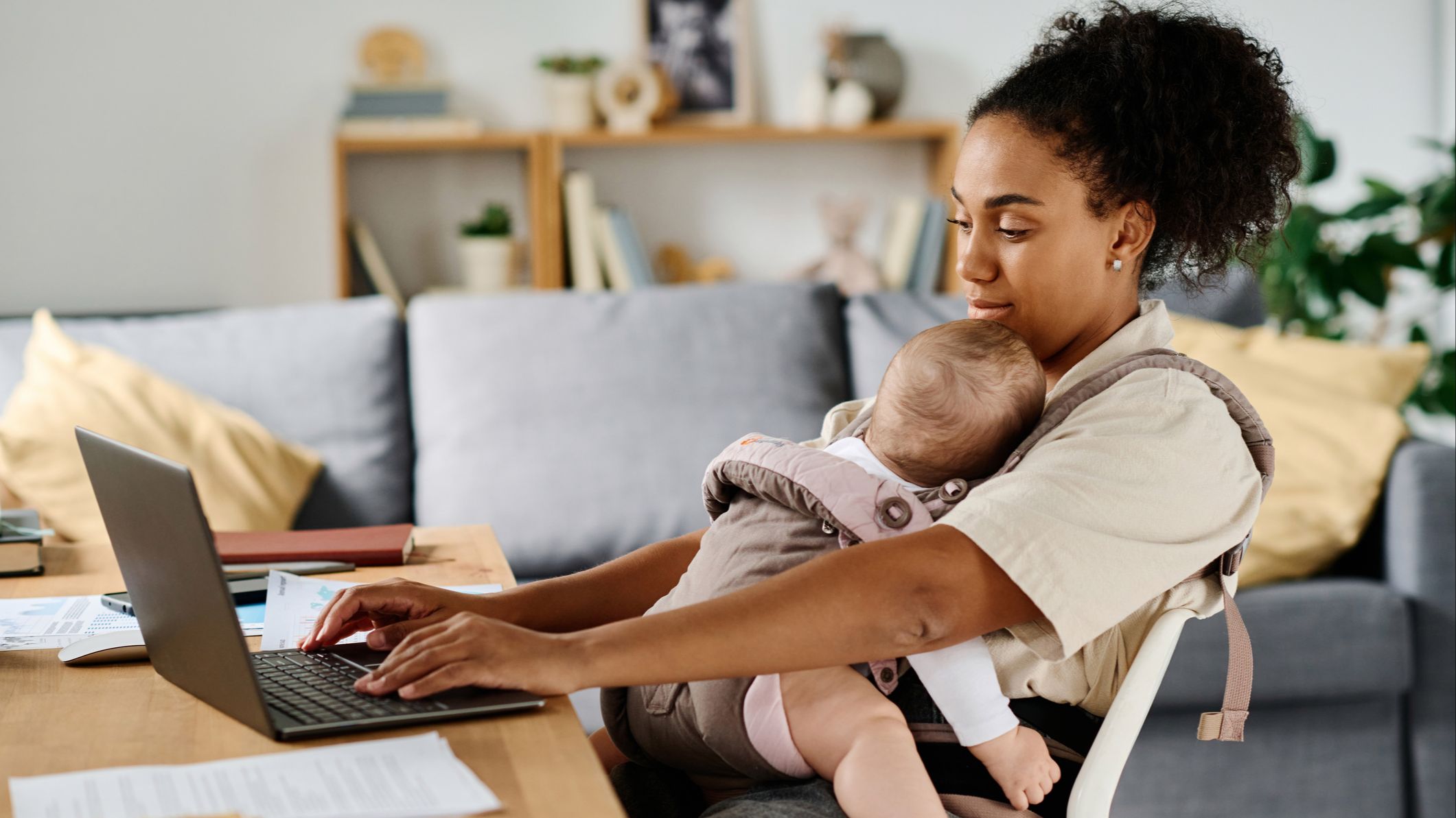 Young mother sitting at table and working online on laptop at home while her baby sleeping in sling