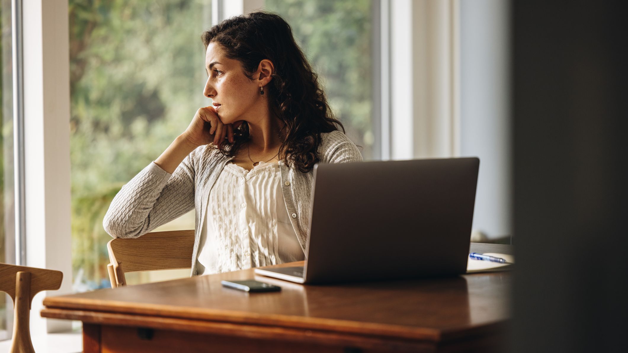 Thoughtful female looking away with hand on chin. Woman sitting at home with laptop on tablet looking away and thinking.
