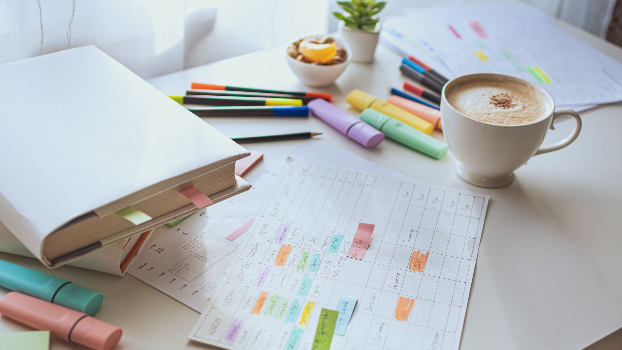 A white table with scattered colorful markers, a weekly schedule and a cup of coffee.