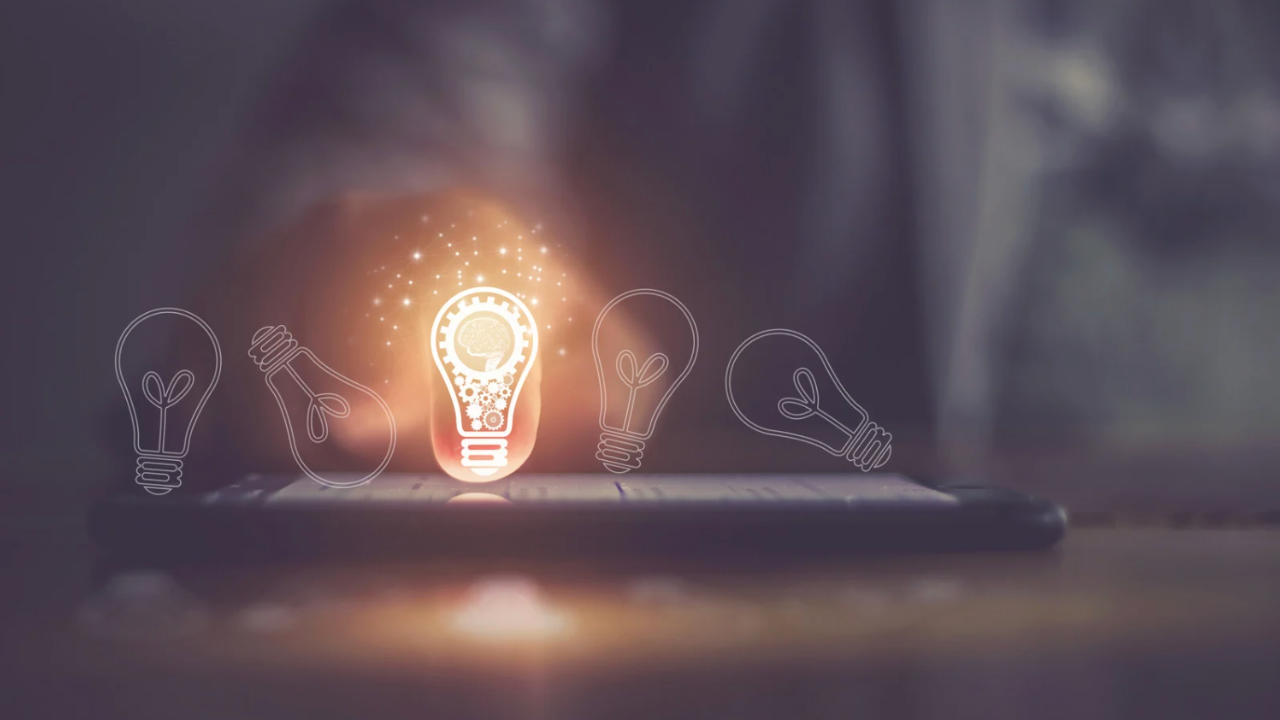 A male instructor stands at the front of a classroom, smiling as he acknowledges a student who has raised their hand to ask a question. The room is filled with students at desks with laptops, creating an engaging and collaborative learning environment.