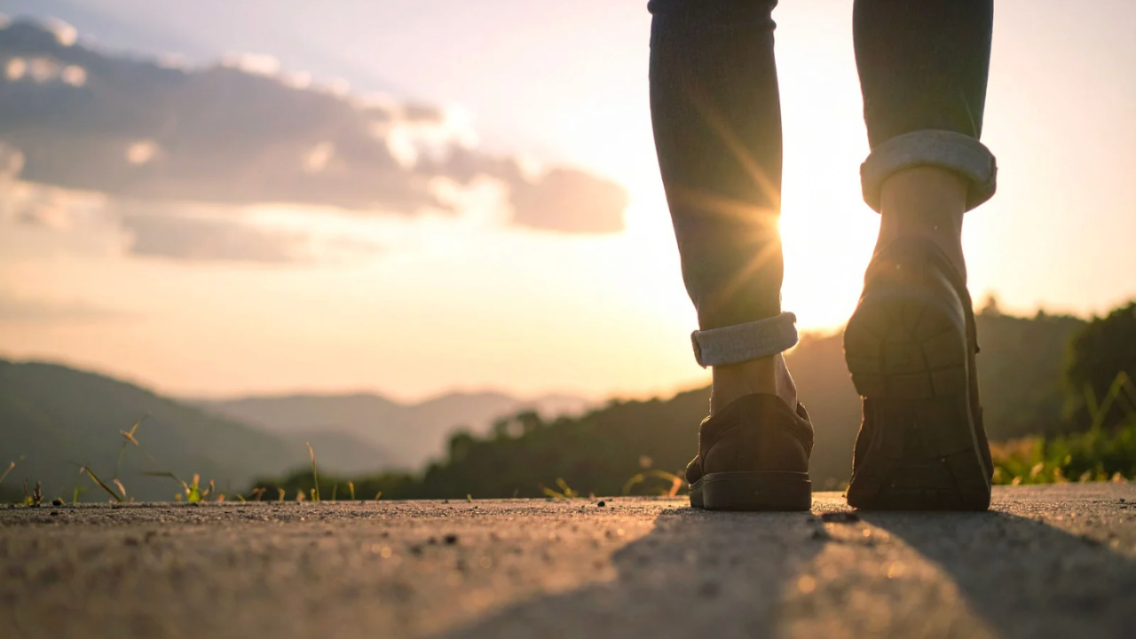A hand is placing a coin on top of the tallest stack in a row of four coin stacks that descend in height from left to right and the background is soft and blurry with warm glowing circles of golden and green light giving it a feeling of financial growth savings or building wealth over time