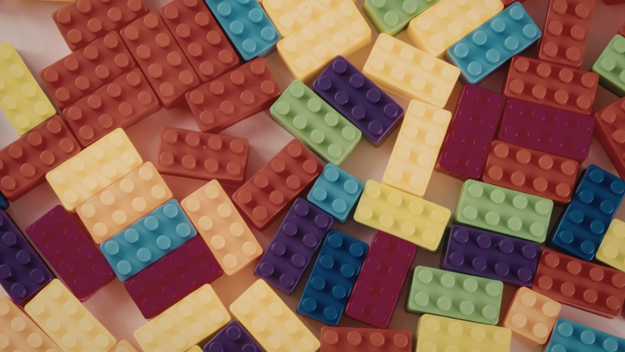 A high-angle shot shows a scattered pile of colorful plastic building blocks resting on a grey baseplate. The primary-colored bricks—red, blue, yellow, and green—are spread out in a disorganized way, suggesting a creative project in progress or a play area.