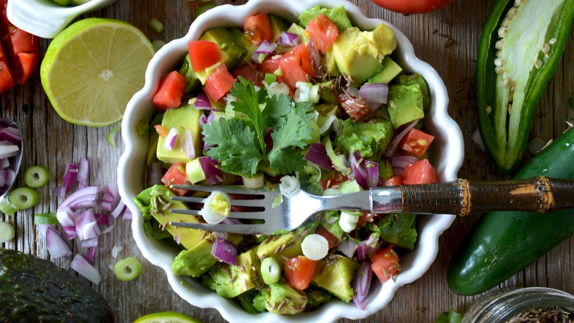 Salad in a white bowl with tomatoes, avocado, cilantro, jalapenos, green onions, red onions, and lime juice all on wood board.