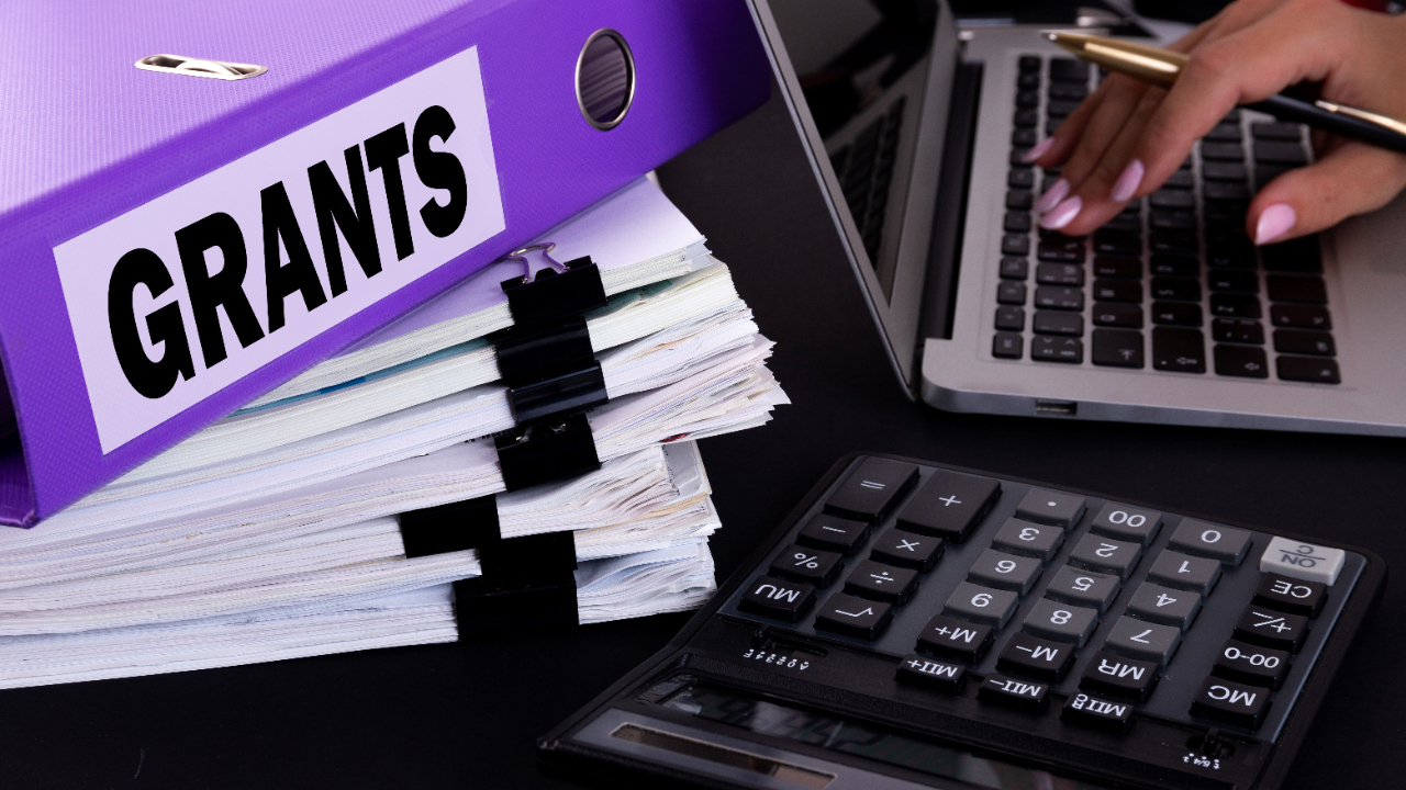 Binder labeled 'Grants,' documents, and a calculator on a desk, illustrating financial planning and answering the Geographic Area of Service question on grant applications for nonprofit success.
