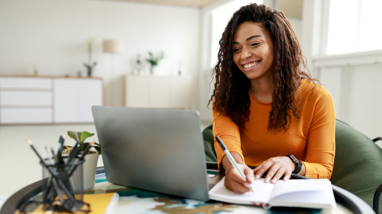 A woman smiling as she writes in a notebook while working on a laptop, representing the process of crafting effective grant application descriptions.