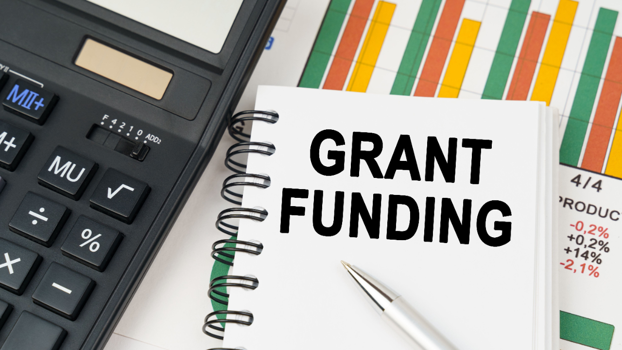 Calculator, financial charts, and a notebook with the words 'Grant Funding' illustrating how to determine the appropriate grant request amount for nonprofit funding success.