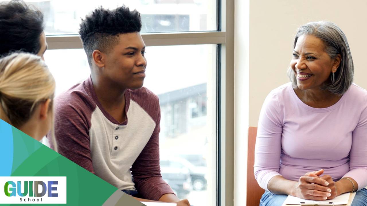 adult woman in pink sweater smiling as she leads a group discussion with teens