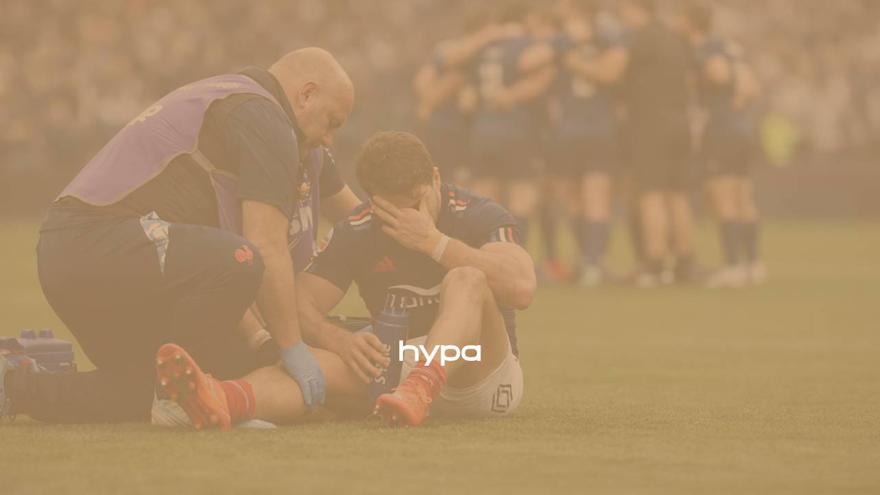 Injured rugby player seated on the field receiving support from coaches while teammates gather in the background during a match.