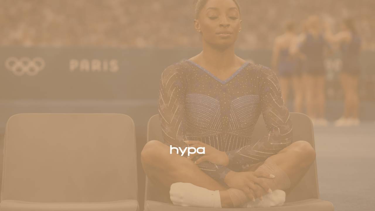 Elite gymnast sits calmly with eyes closed before competition, centred and composed amid a blurred stadium crowd and fellow athletes in the background.