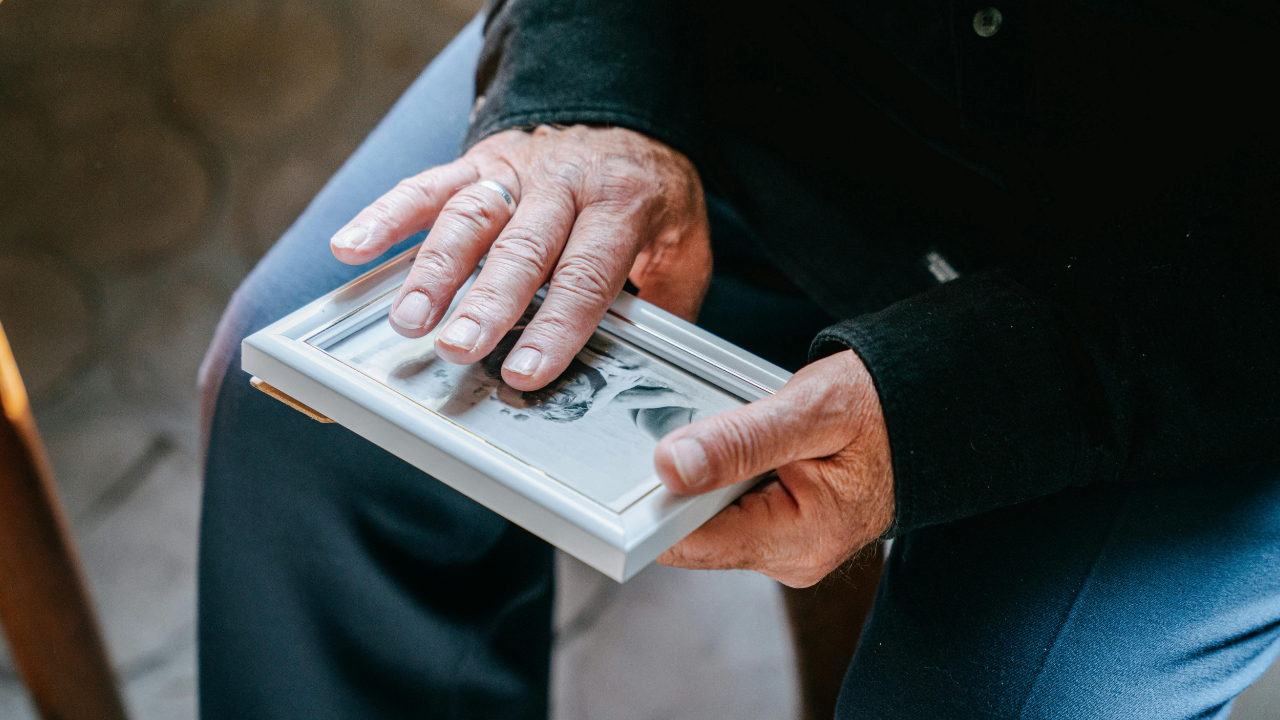 An older person’s hands gently hold a small framed black-and-white photograph while seated indoors.
