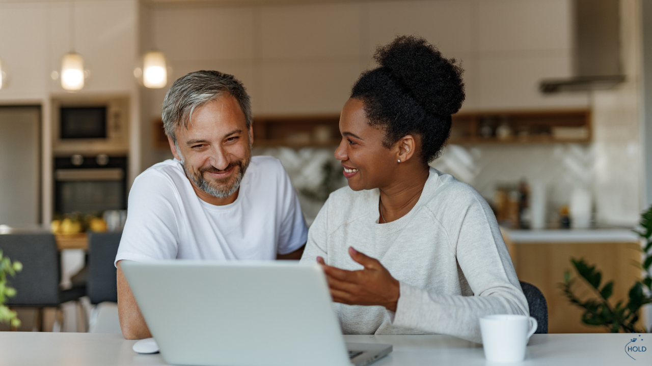 A man and a woman sit at a kitchen table, smiling and talking while looking at a laptop. Their body language suggests trust, connection, and engaged listening, aligning with the blog’s themes of support and healthy communication.