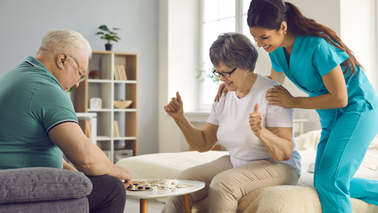 Care worker supporting an older woman and man while they play a board game, showing person-centred care, encouragement, and positive engagement in a home setting.