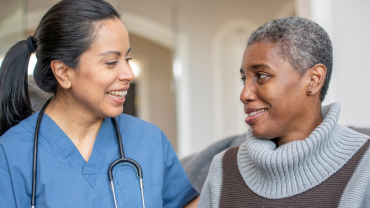 A nurse wearing blue scrubs and a stethoscope smiles warmly at an older woman, who is sitting and smiling back.