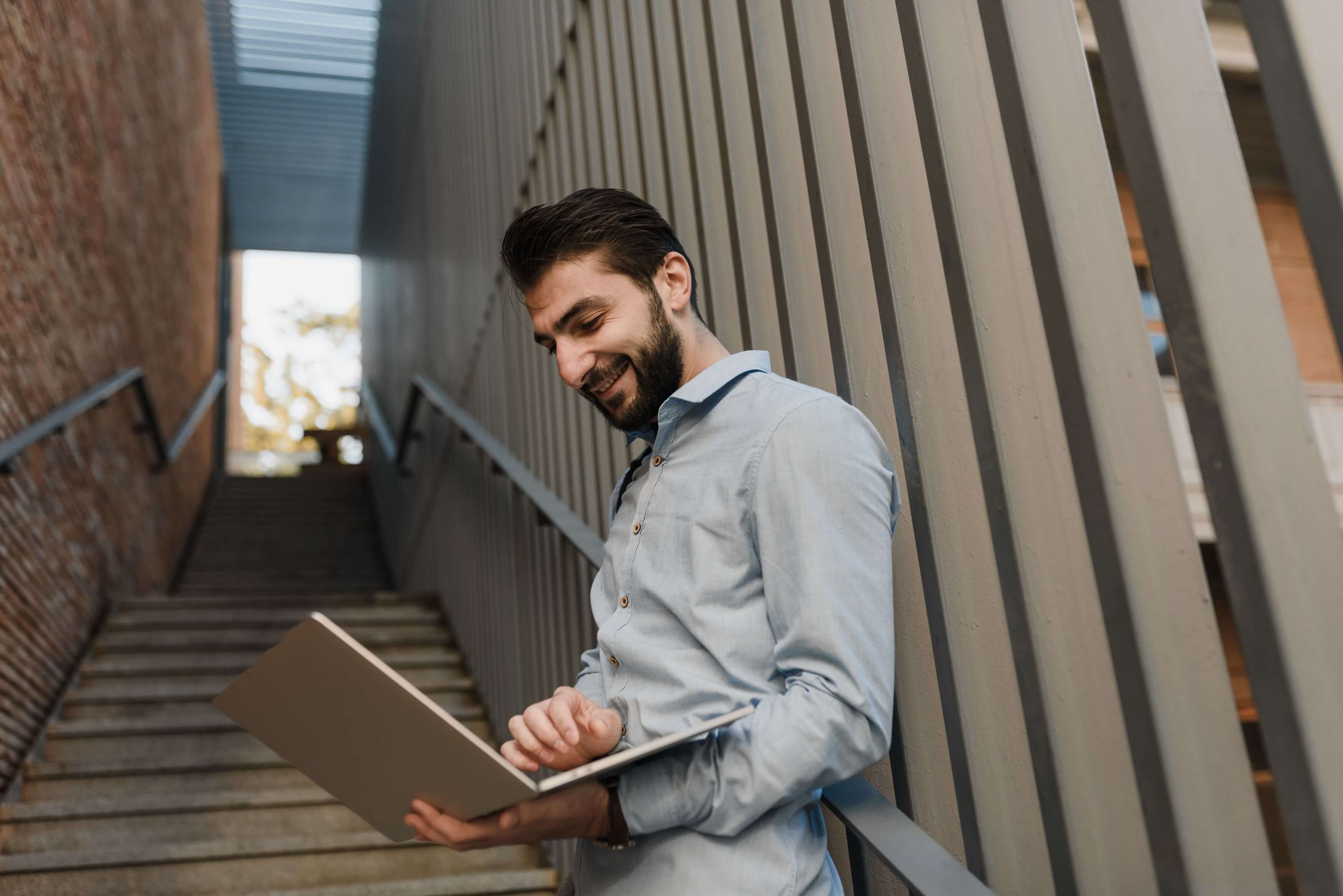 Bearded man in a light blue shirt smiling down at a laptop while leaning against a metal wall beside an outdoor staircase | therapeutic coaching online | international trauma therapist | executive coaching | Los Angeles | Seattle | New York City