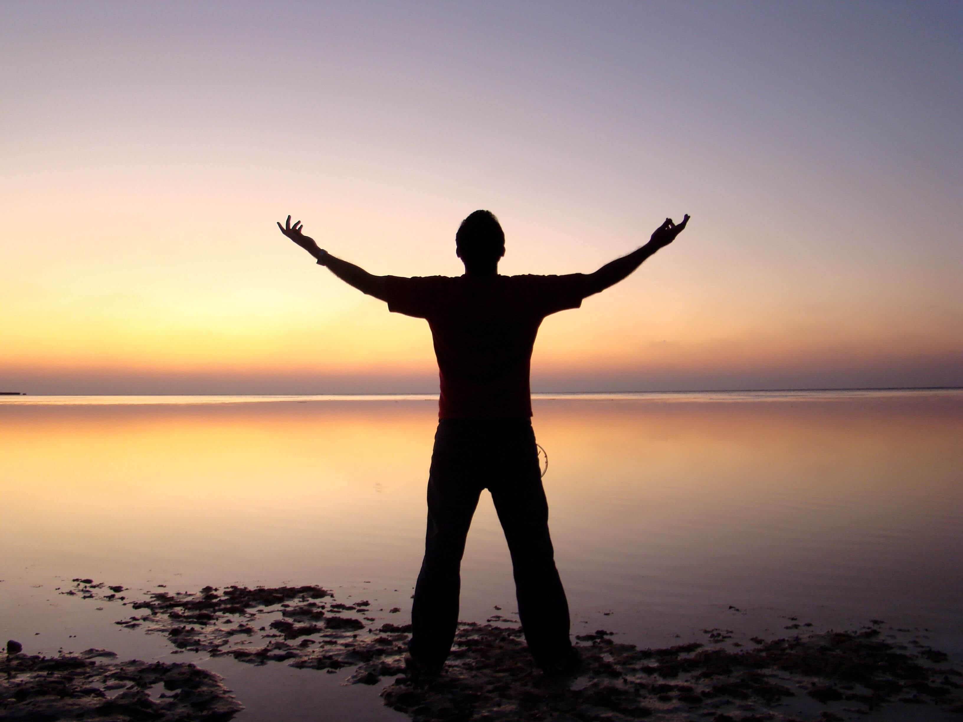 Silhouette of a man standing with arms stretched wide open facing a calm reflective ocean at sunset in warm golden light | energy healing trauma | therapeutic coaching online | international trauma therapist | Seattle | Los Angeles | New York City
