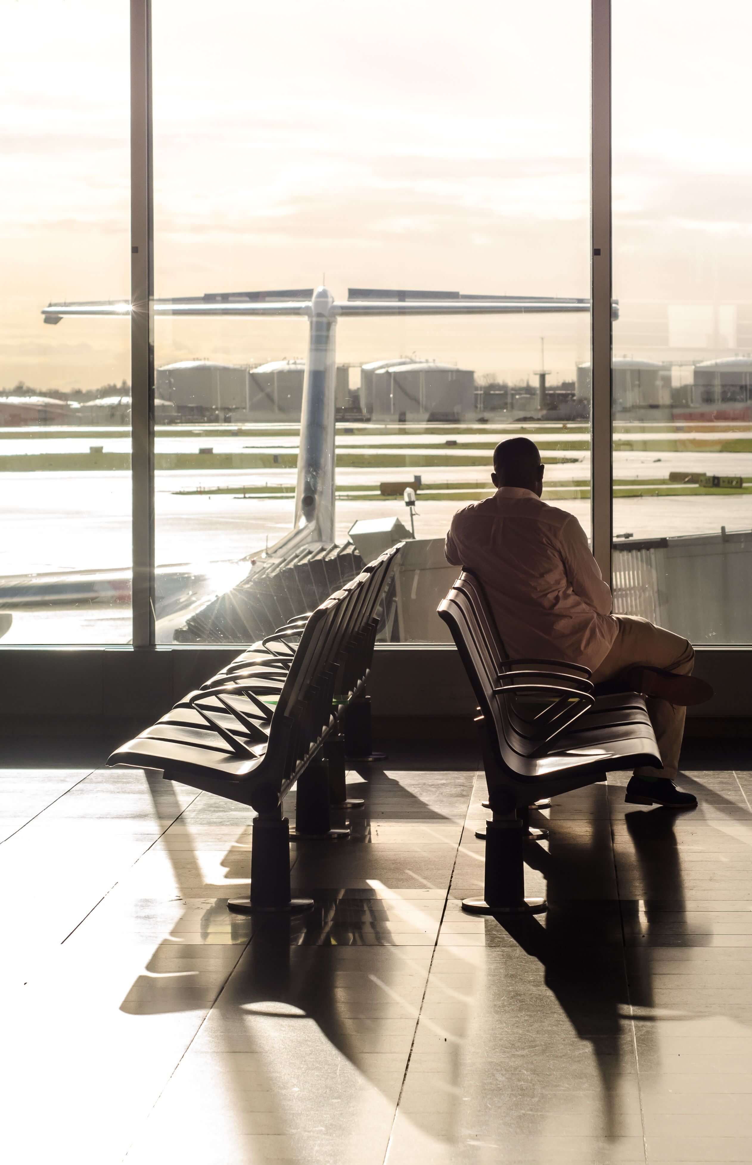 Man sitting alone on airport seating looking out through large windows at a plane on the runway in golden light | expat community | deep connection expat | international therapy and coaching | England | Netherlands | Germany