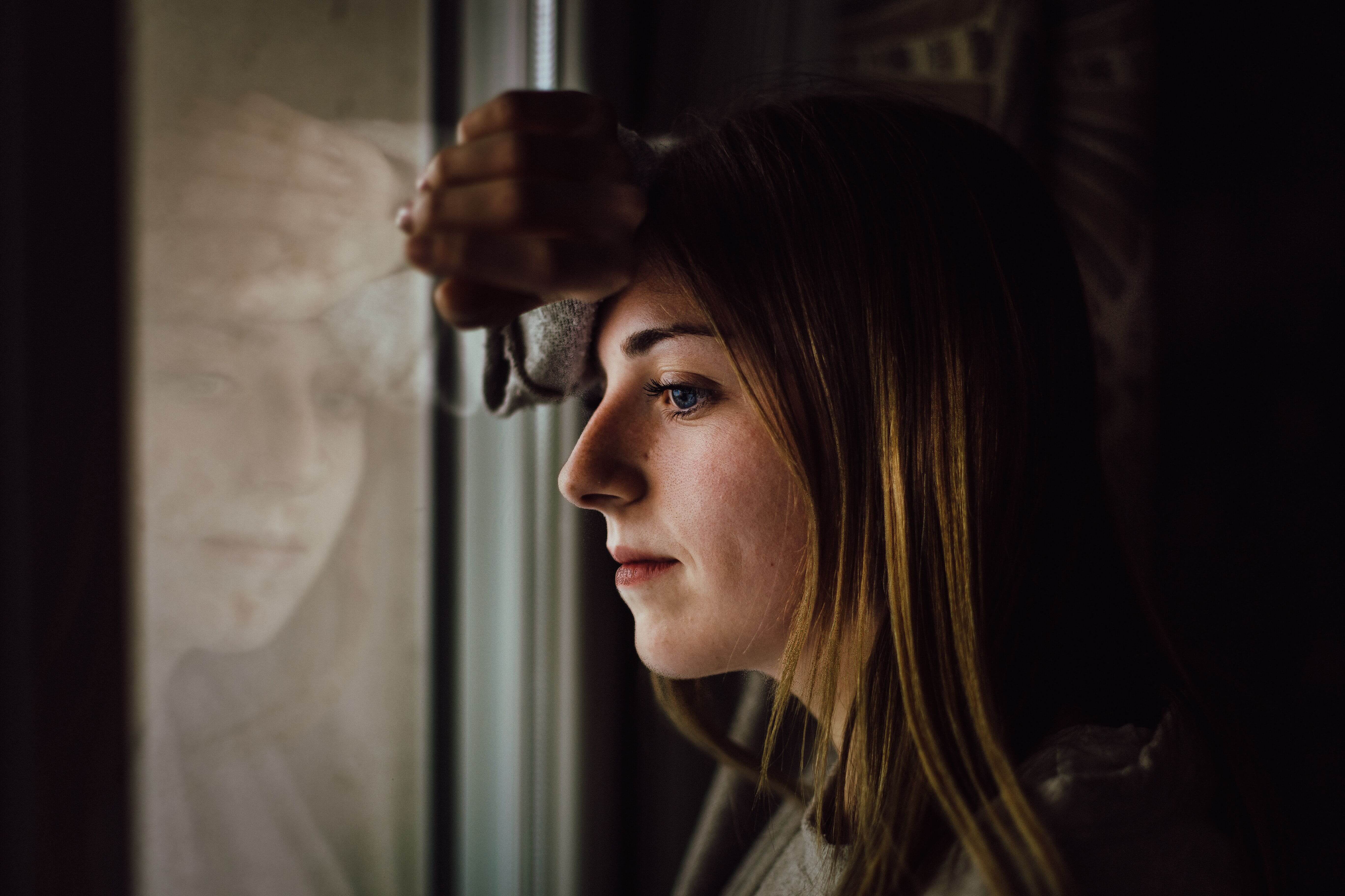 Young woman with blonde hair leaning against a dark window frame looking pensively into the distance with her reflection visible | tapping therapy online | energy psychology online | international trauma therapist | Chicago | New York City | Seattle
