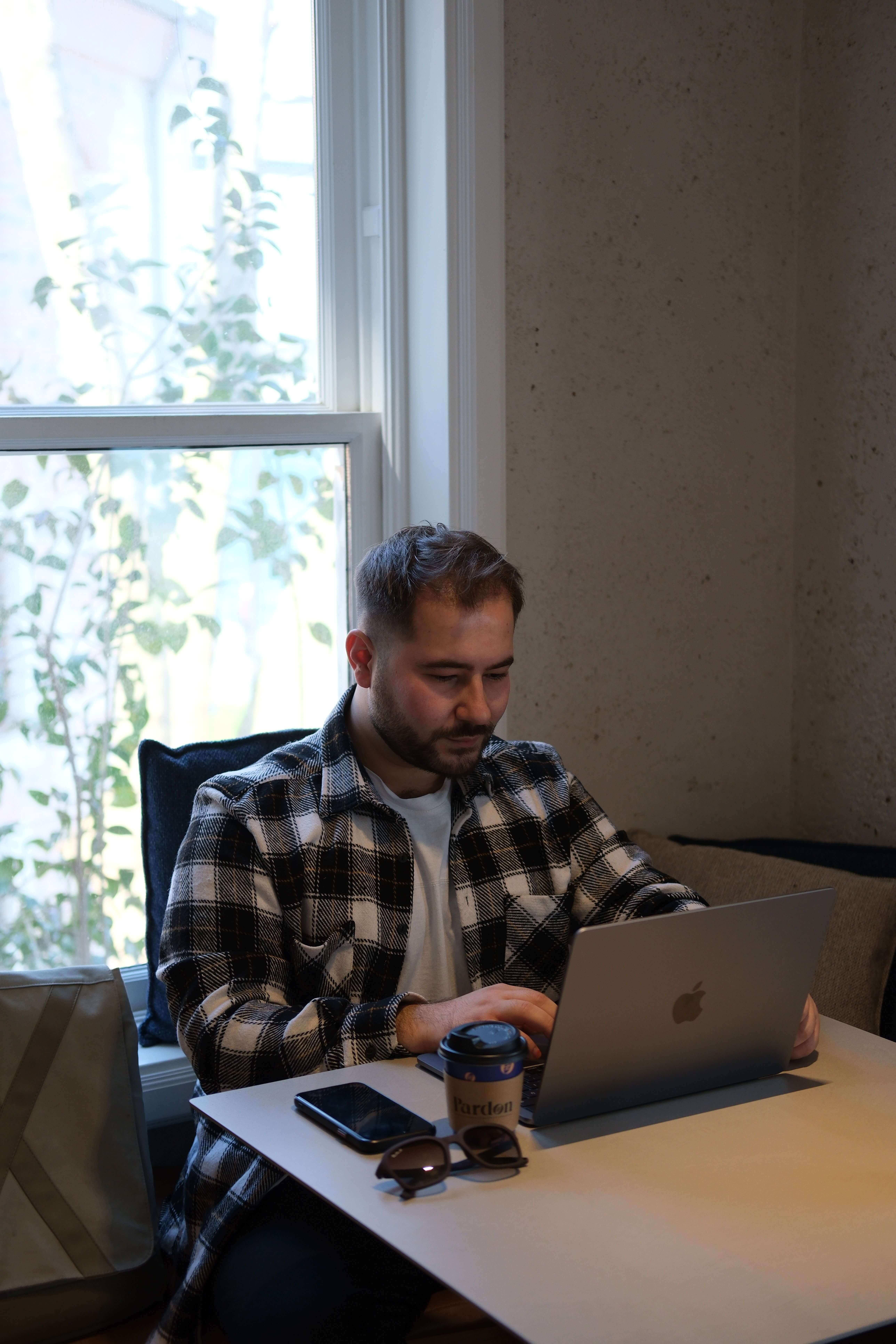 Man in a black and white plaid shirt working on a MacBook at a white table by a bright window with a coffee cup and sunglasses beside him | parts work training for therapists | executive coaching | international trauma therapist | Los Angeles | Seattle | Chicago