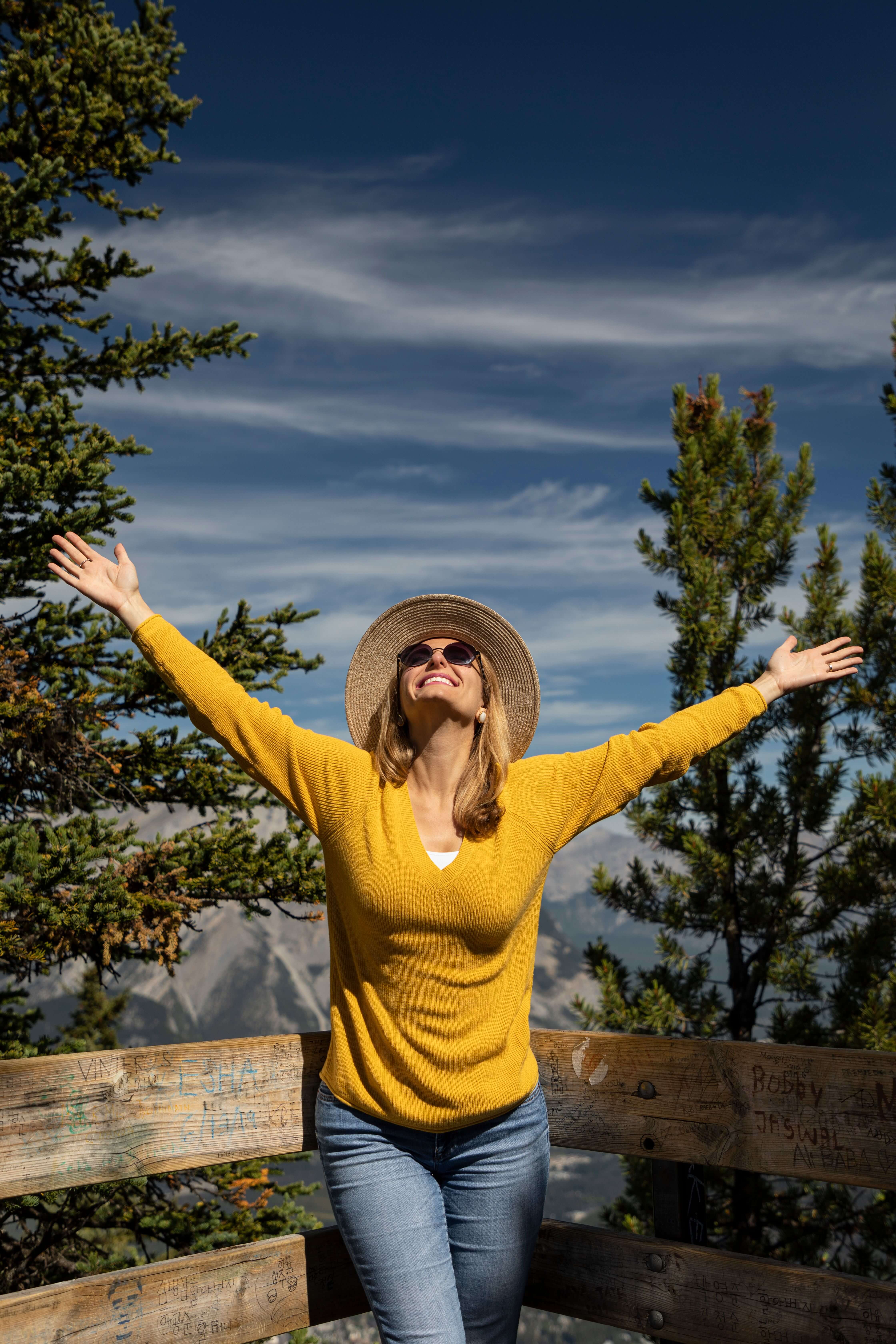 Woman in a yellow sweater and sun hat smiling with arms stretched wide open standing on a mountain viewpoint with pine trees behind her | therapeutic coaching online | private pay therapy alternative | international trauma therapist | Seattle | Los Angeles | New York City