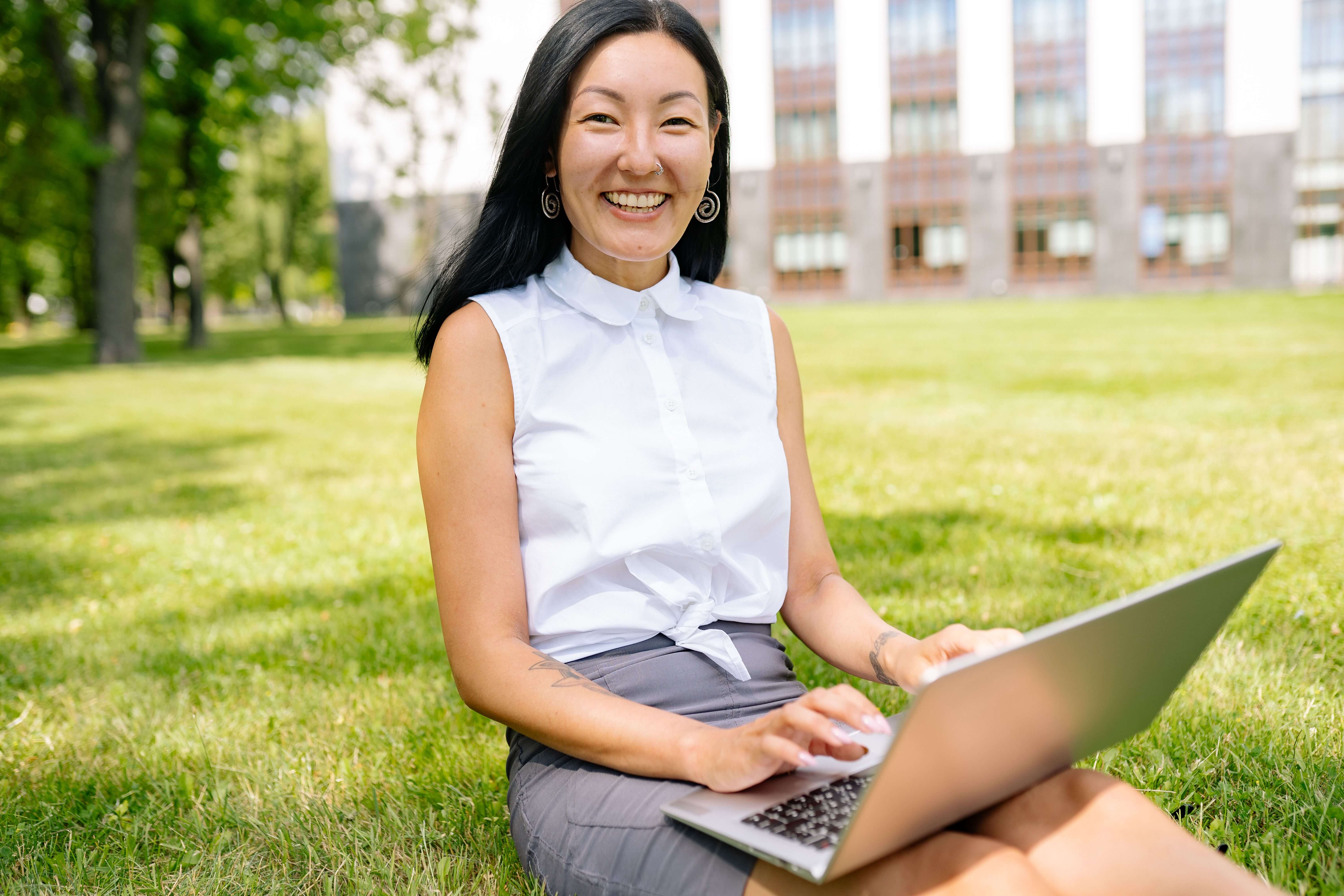 Woman with long dark hair and a nose ring smiling at the camera while sitting on grass with a laptop in a sunny outdoor setting | international therapist | therapeutic coaching | executive coaching | Seattle | New York City | Chicago