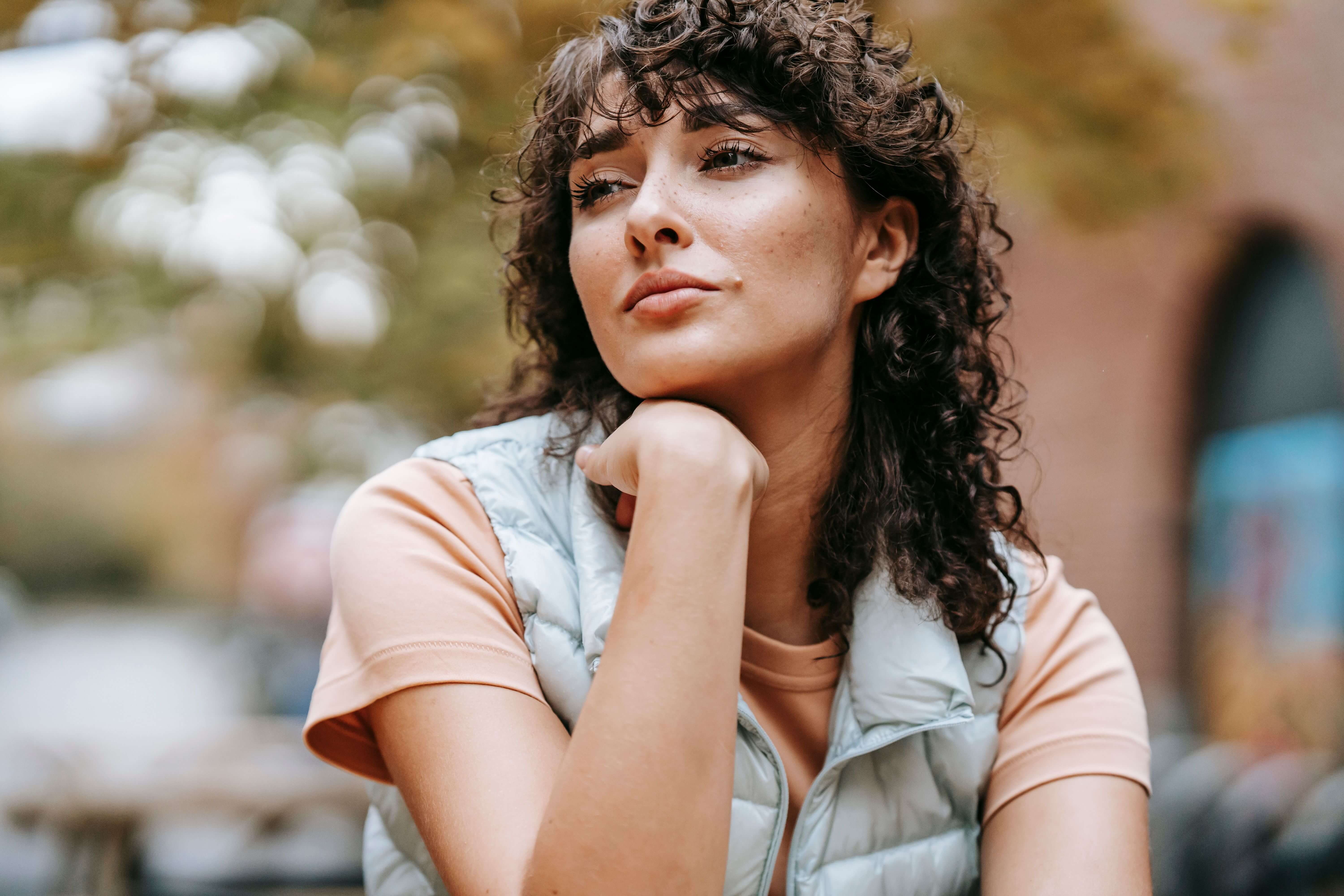 Young woman with curly dark hair and a puffer vest resting her chin on her fist looking pensively into the distance outdoors | therapy vs coaching | therapeutic coaching | international trauma therapist | Chicago | New York City | Seattle