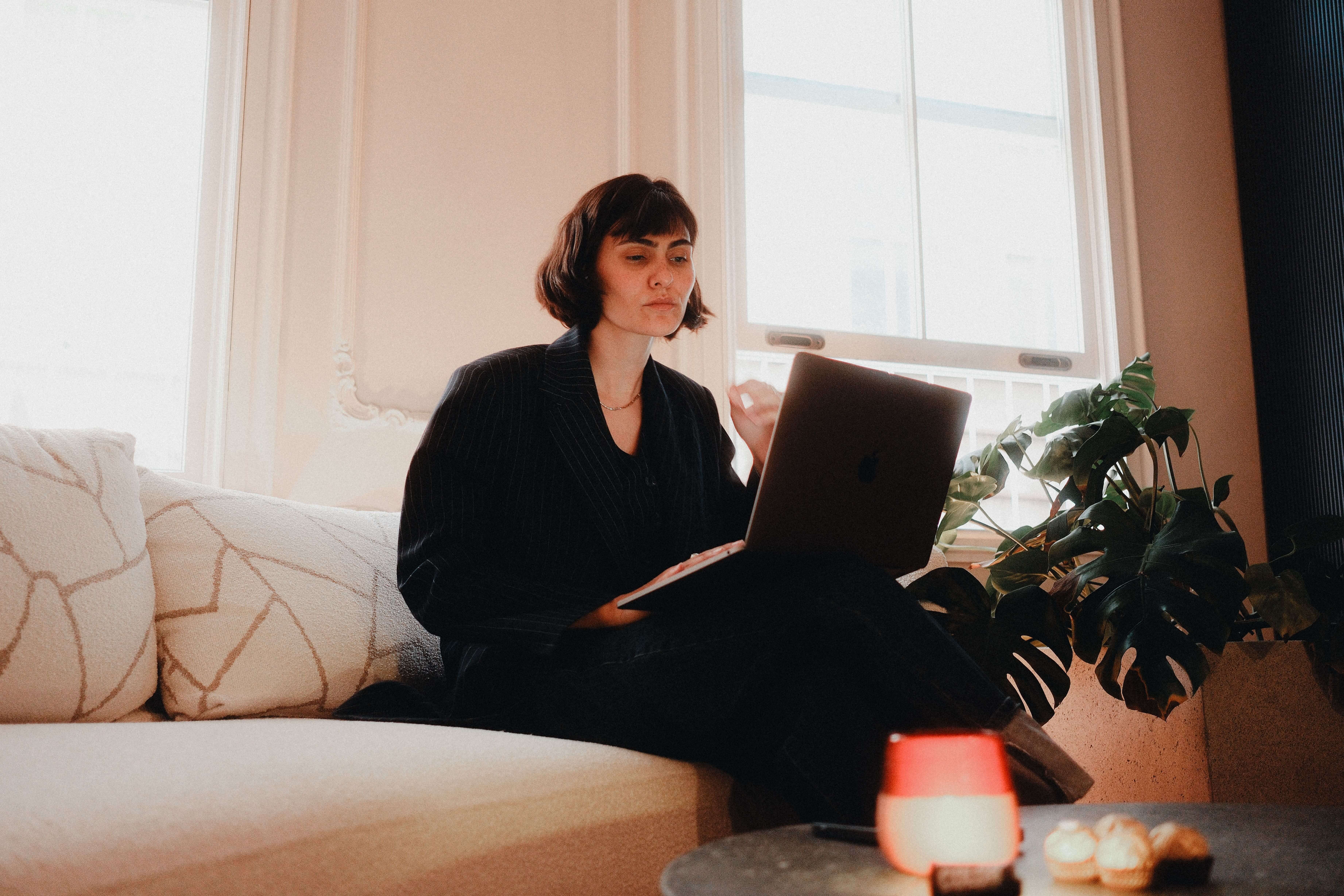 Woman with a short dark bob sitting cross legged on a white sofa with a laptop and notebook in a bright room with large windows and a monstera plant | therapeutic coaching online | parts work training | international trauma therapist | New York City | Los Angeles | Seattle