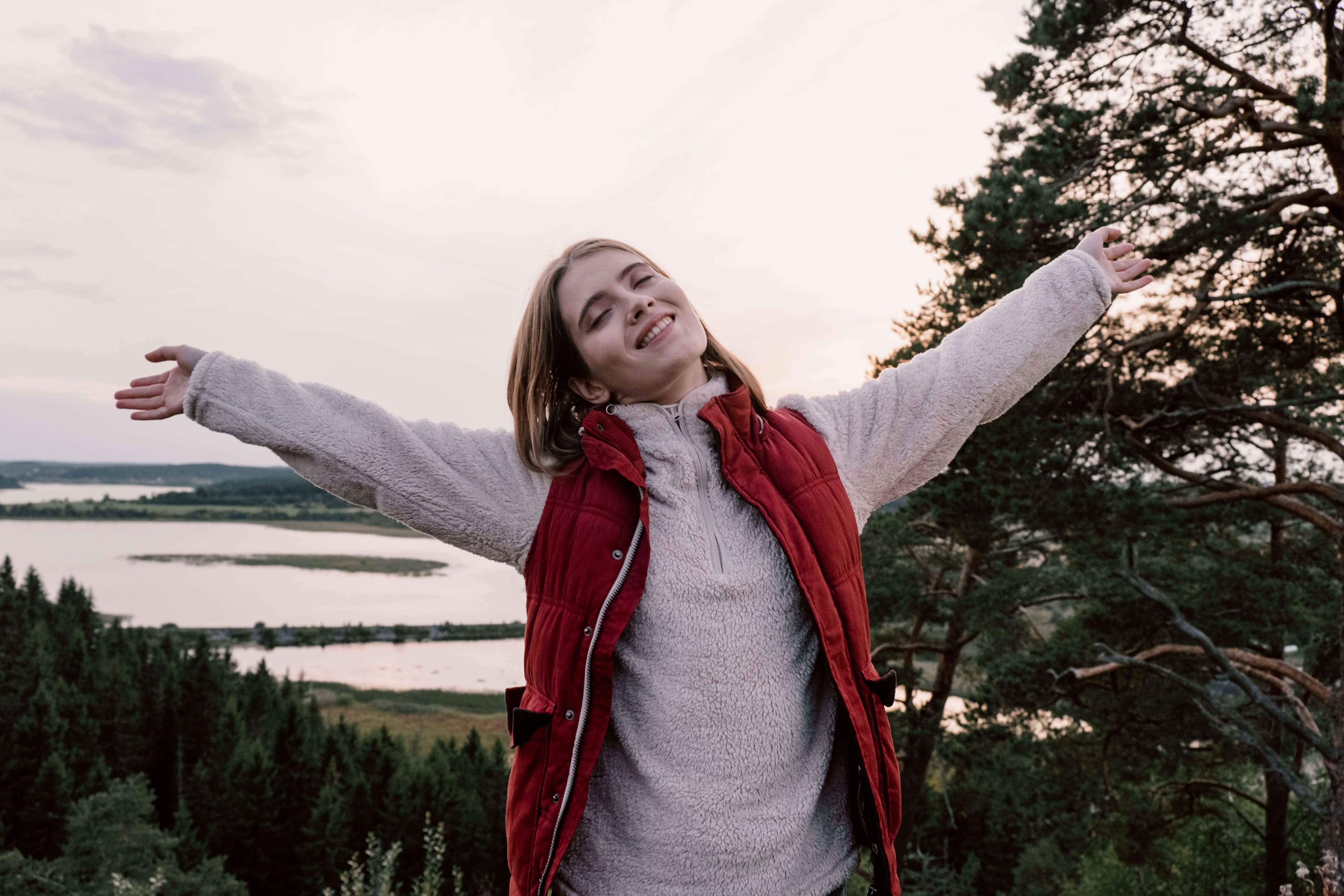 Young woman in a red gilet smiling with arms stretched wide open standing on a hilltop overlooking a lake and forest | therapeutic coaching | private pay therapy alternative | international trauma therapist | Chicago | New York City | Seattle