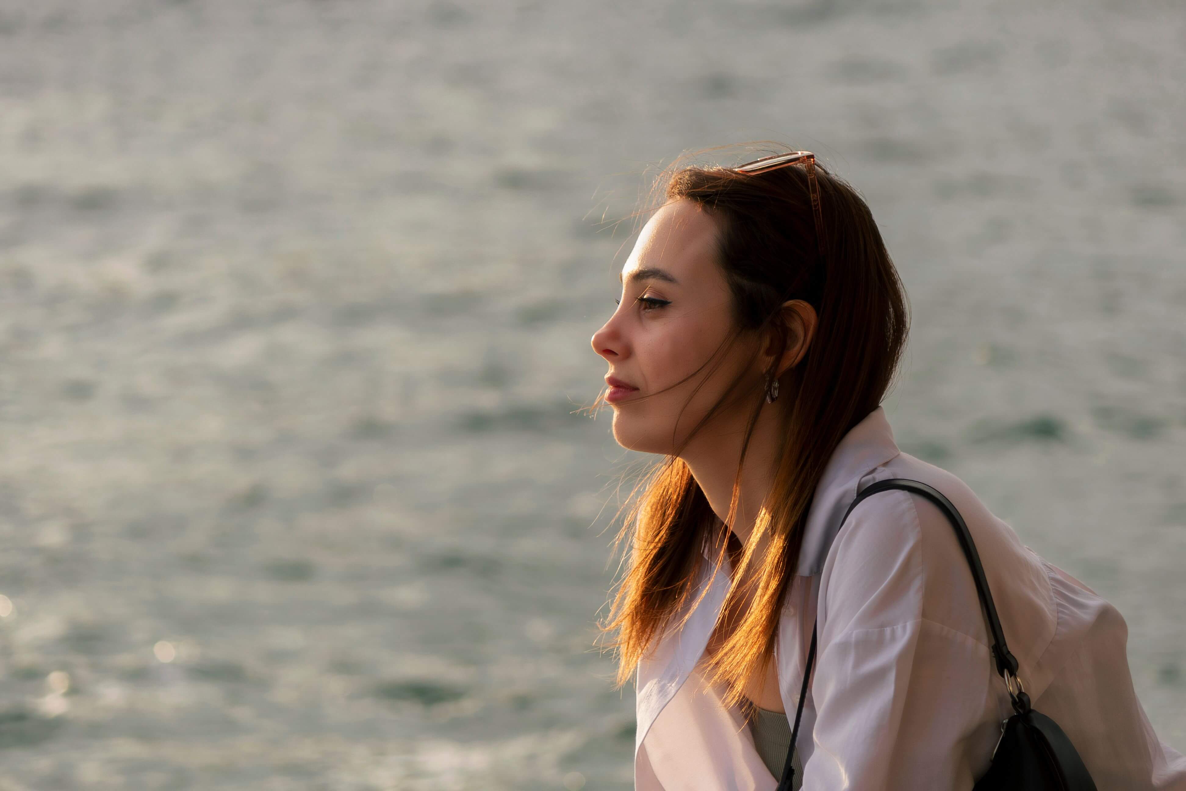 Woman with auburn hair and sunglasses on her head standing in profile against a shimmering body of water in warm golden light | therapeutic coaching online | international trauma therapist | parts work training for therapists | New York City | Chicago | Los Angeles