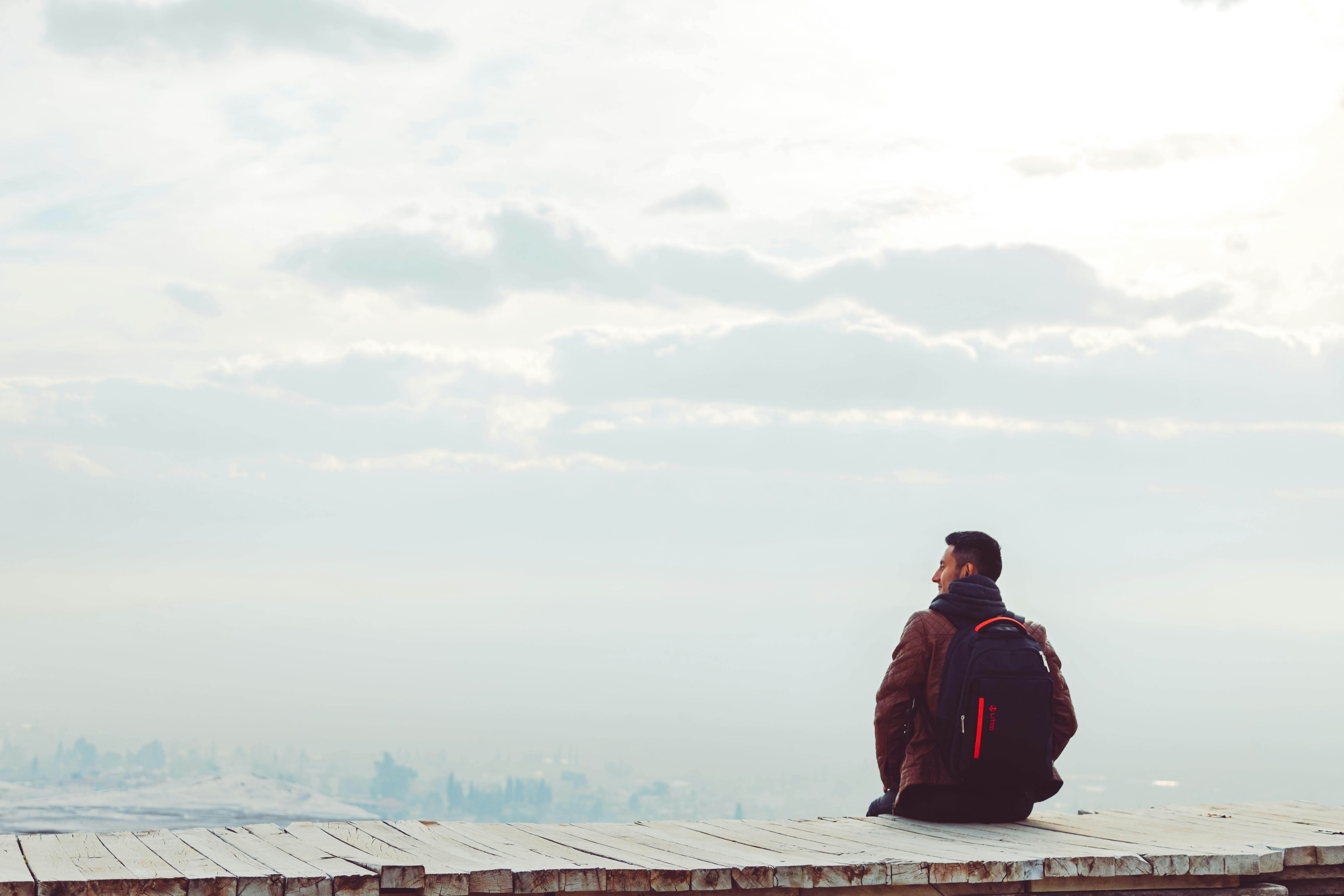 Man with a backpack sitting alone on a wooden platform overlooking a hazy city skyline and cloudy sky | therapy across borders | international therapist | expat mental health support | Portugal | South Africa | Italy