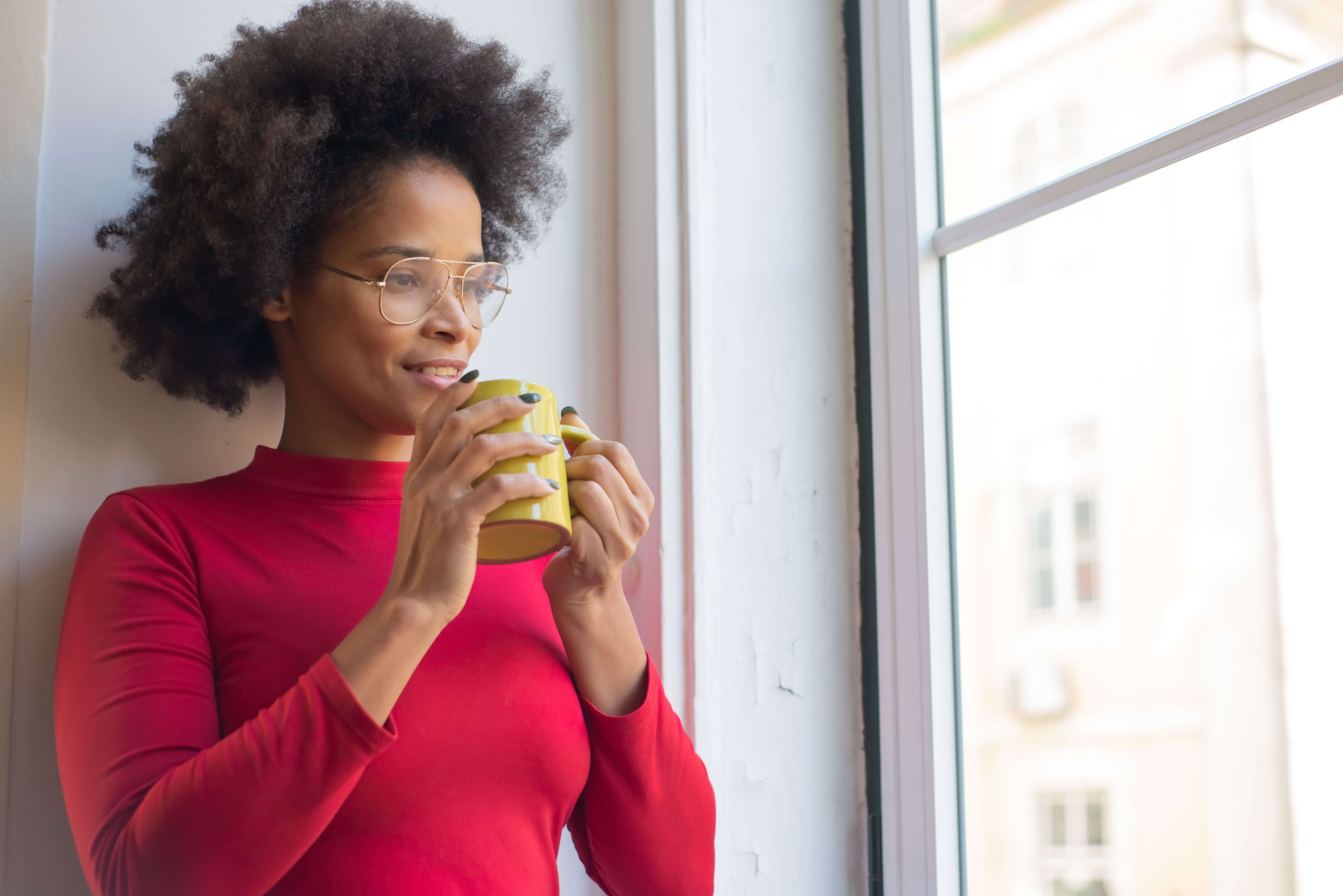 Woman with natural hair and glasses holding a yellow mug looking thoughtfully out of a bright window | therapy for expats | expat identity | international therapist | United States | England | Netherlands