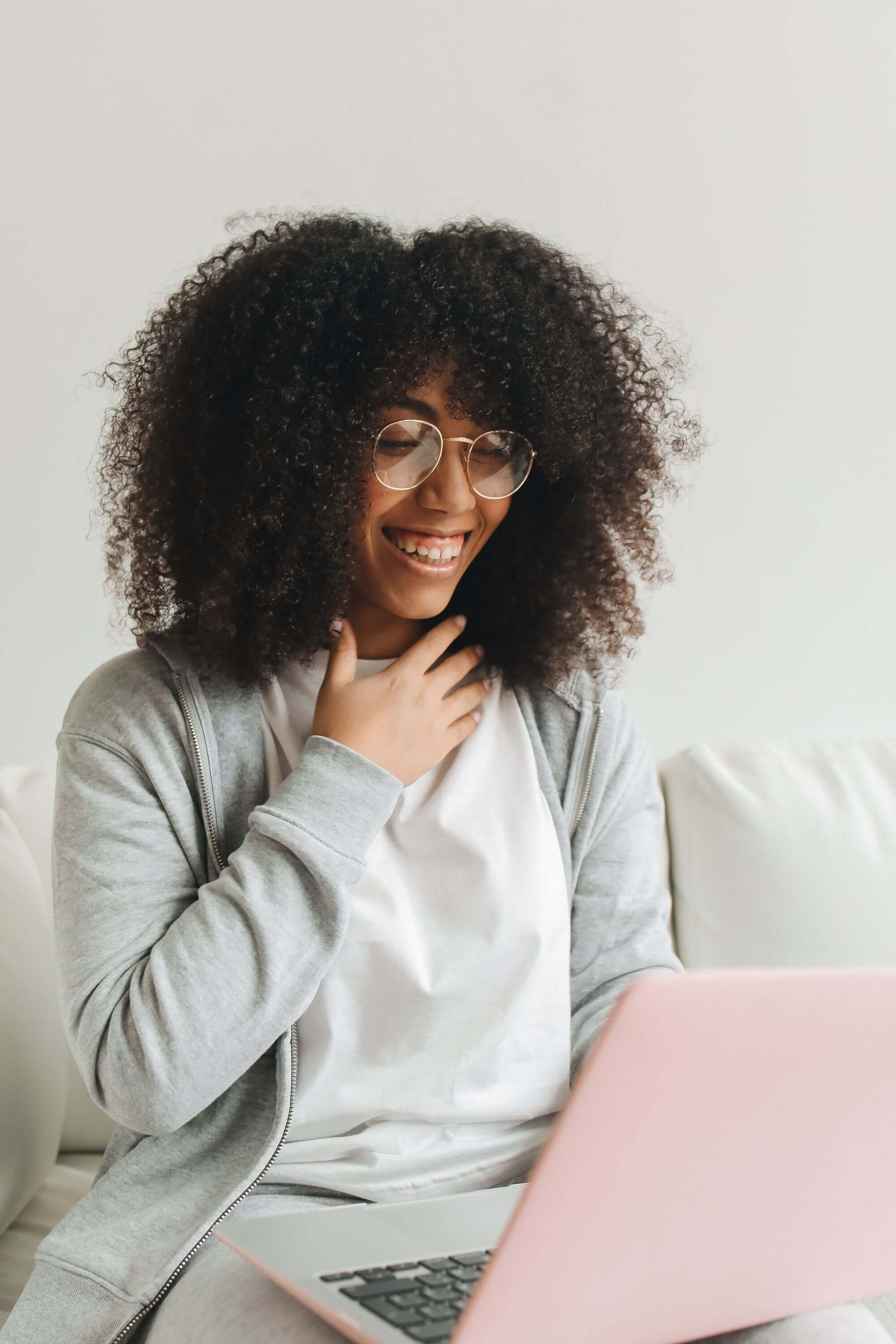 Woman with large curly hair and glasses laughing and touching her chest while looking at a pink laptop on a white sofa | therapeutic coaching online | international trauma therapist | single session coaching online | New York City | Chicago | Los Angeles