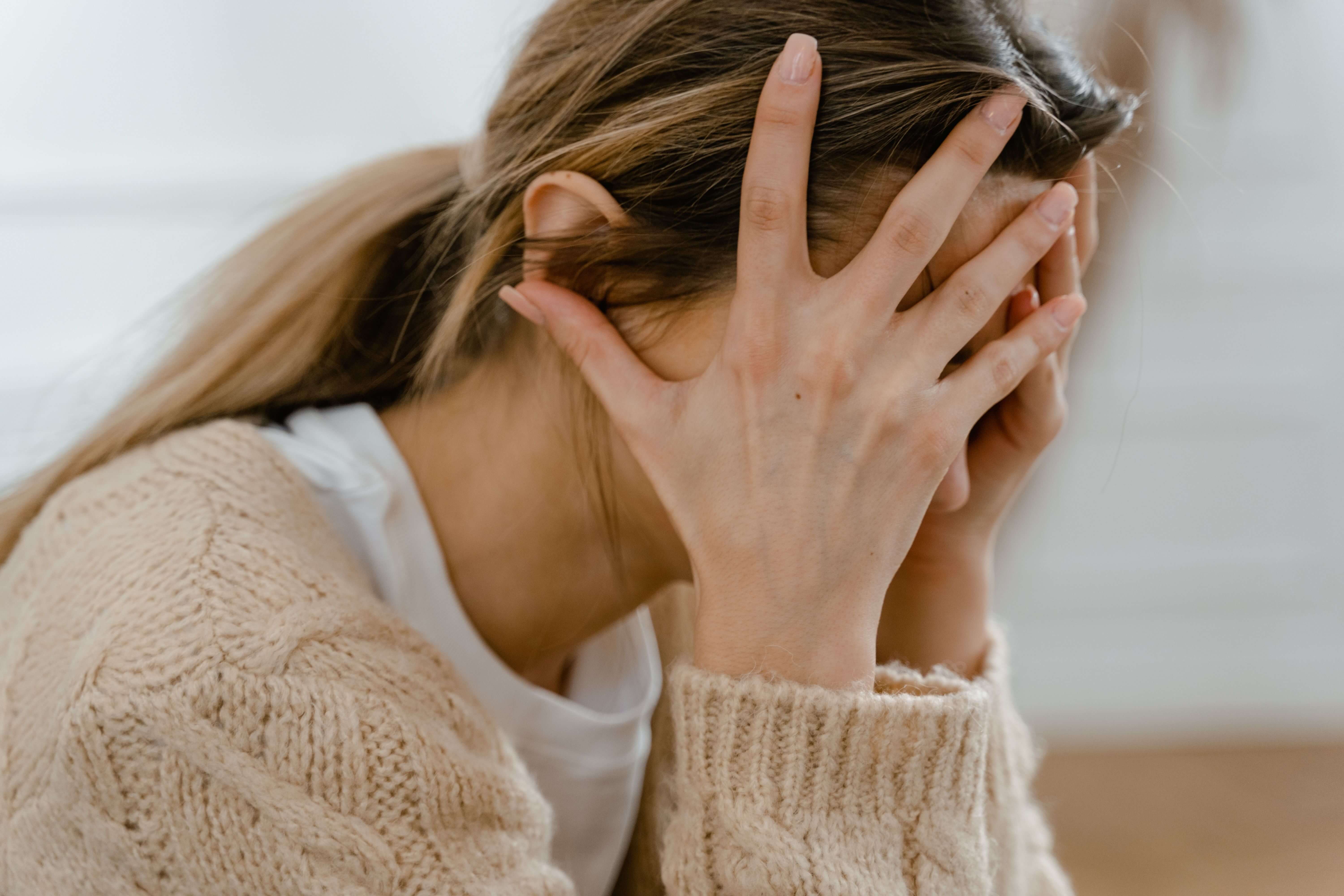 Woman in a beige knit cardigan sitting with her head in her hands in a bright white room | high functioning trauma | burnout trauma professionals | international trauma therapist | England | Netherlands | Germany