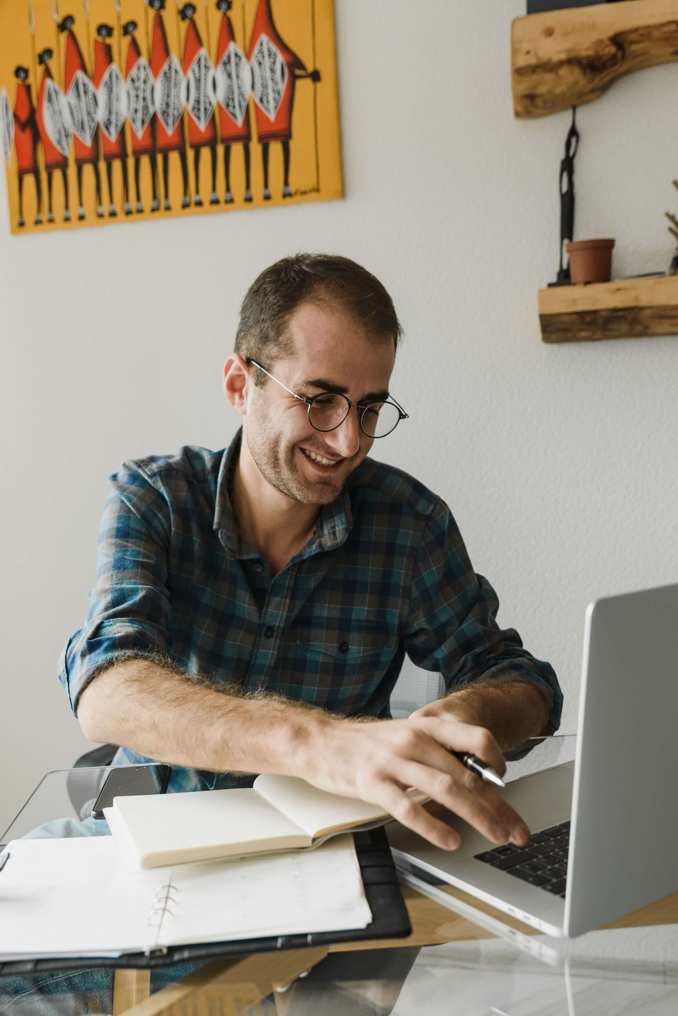 Man with round glasses and a plaid shirt smiling while writing in a notebook at a glass desk with a laptop and colourful artwork on the wall behind him | parts work training for therapists | international trauma therapist | therapeutic coaching | Chicago | New York City