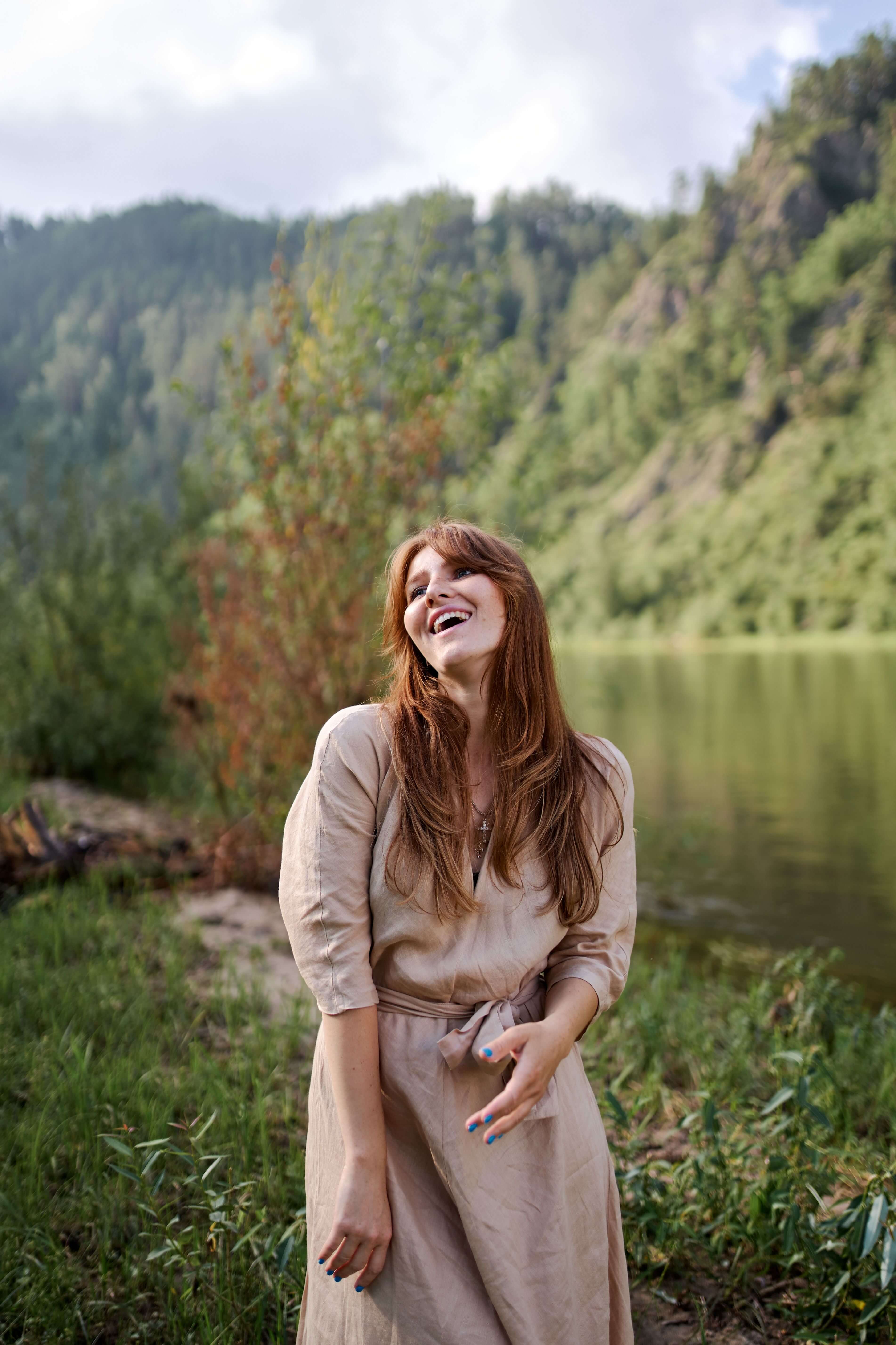 Woman with long red hair laughing with her head back standing beside a river with forested hills behind her | trauma therapy for high achievers | EMDR therapy online | international trauma therapist | Scotland | Canada | France