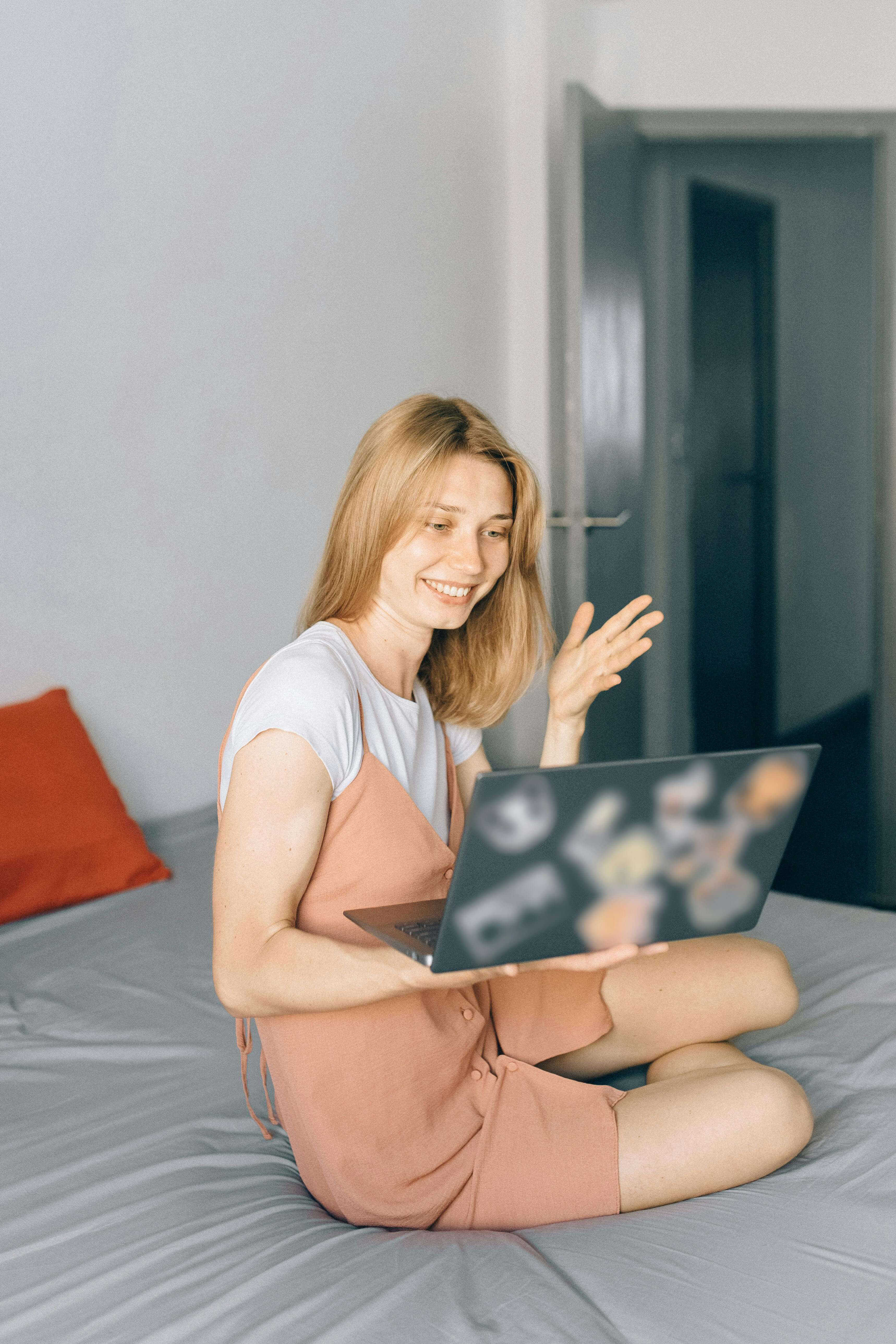 Young woman sitting cross-legged on a bed smiling and waving at a laptop screen during an online session | international therapist | digital nomad mental health | United States | Germany