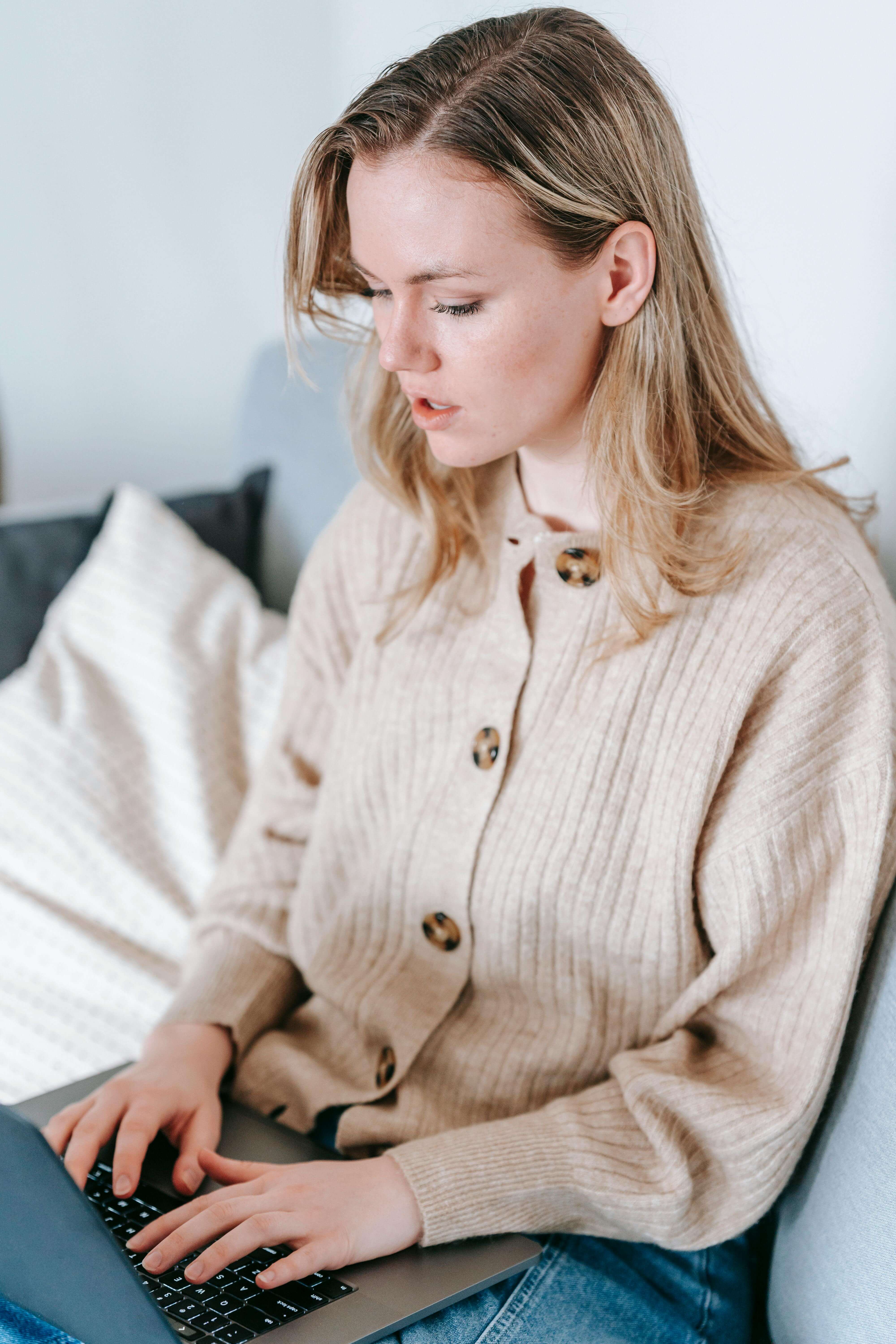 Young blonde woman in a beige cardigan sitting on a sofa looking intently at a laptop screen | international therapist | expat intimacy avoidance | therapy for expats | United States | Canada | Portugal