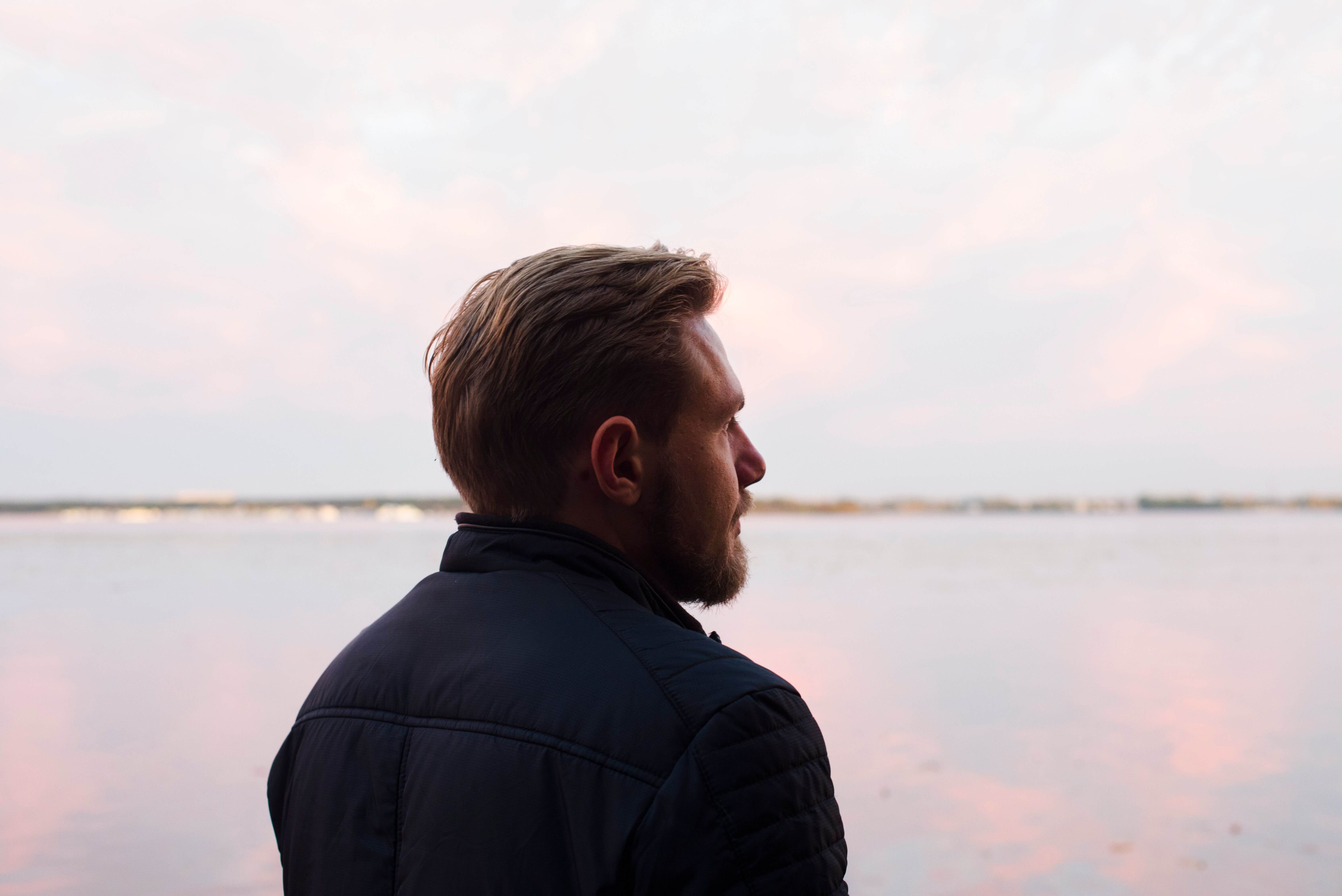 Man with a beard in a dark jacket standing with his back to the camera looking out over a calm pink tinted lake at dusk | what is therapeutic coaching | therapeutic coaching online | international trauma therapist | Seattle | Los Angeles | New York City