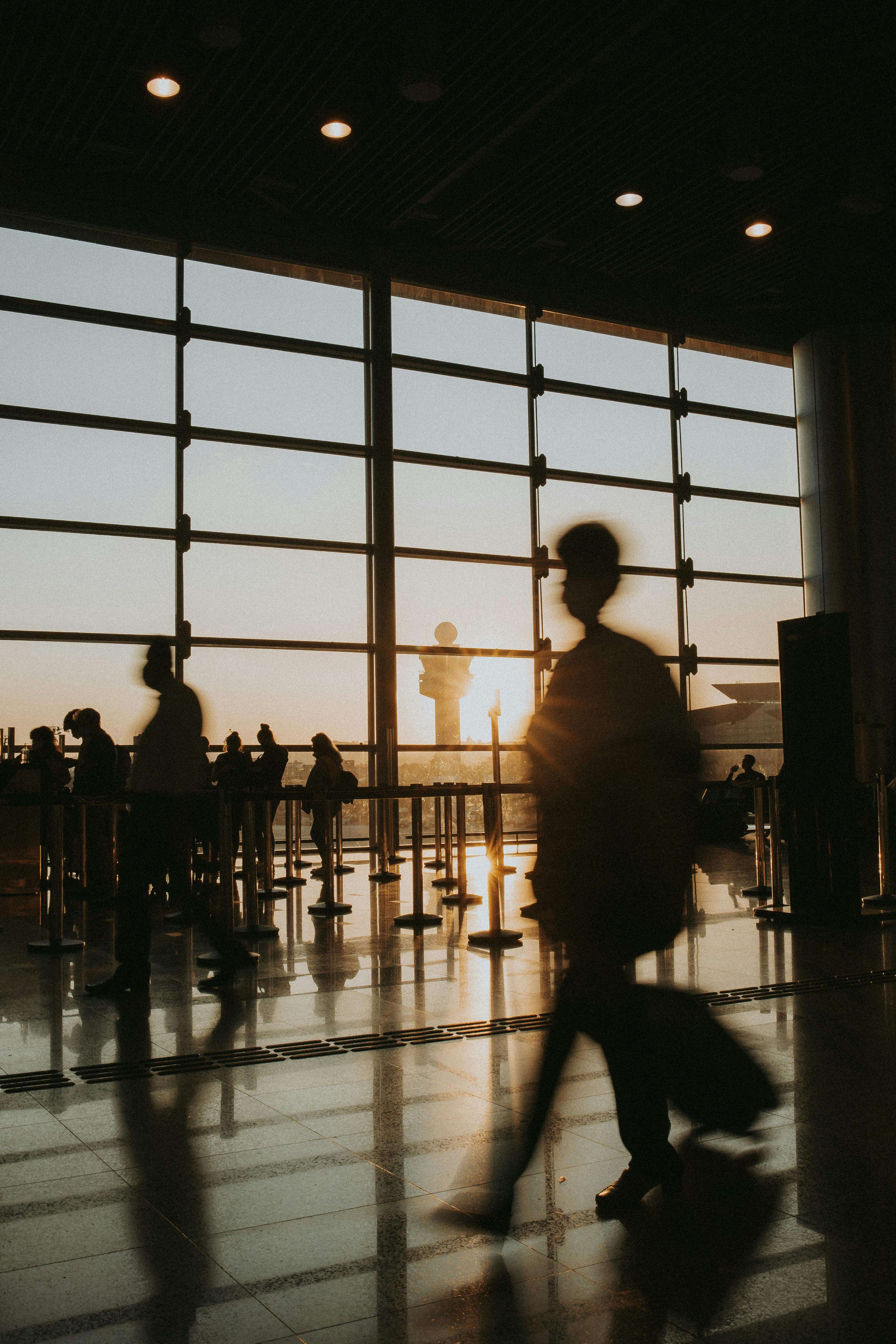 Silhouette of a traveller walking through a busy airport terminal at sunset with a rolling suitcase | expat burnout | aid worker burnout | international therapy and coaching | Portugal | France | Italy