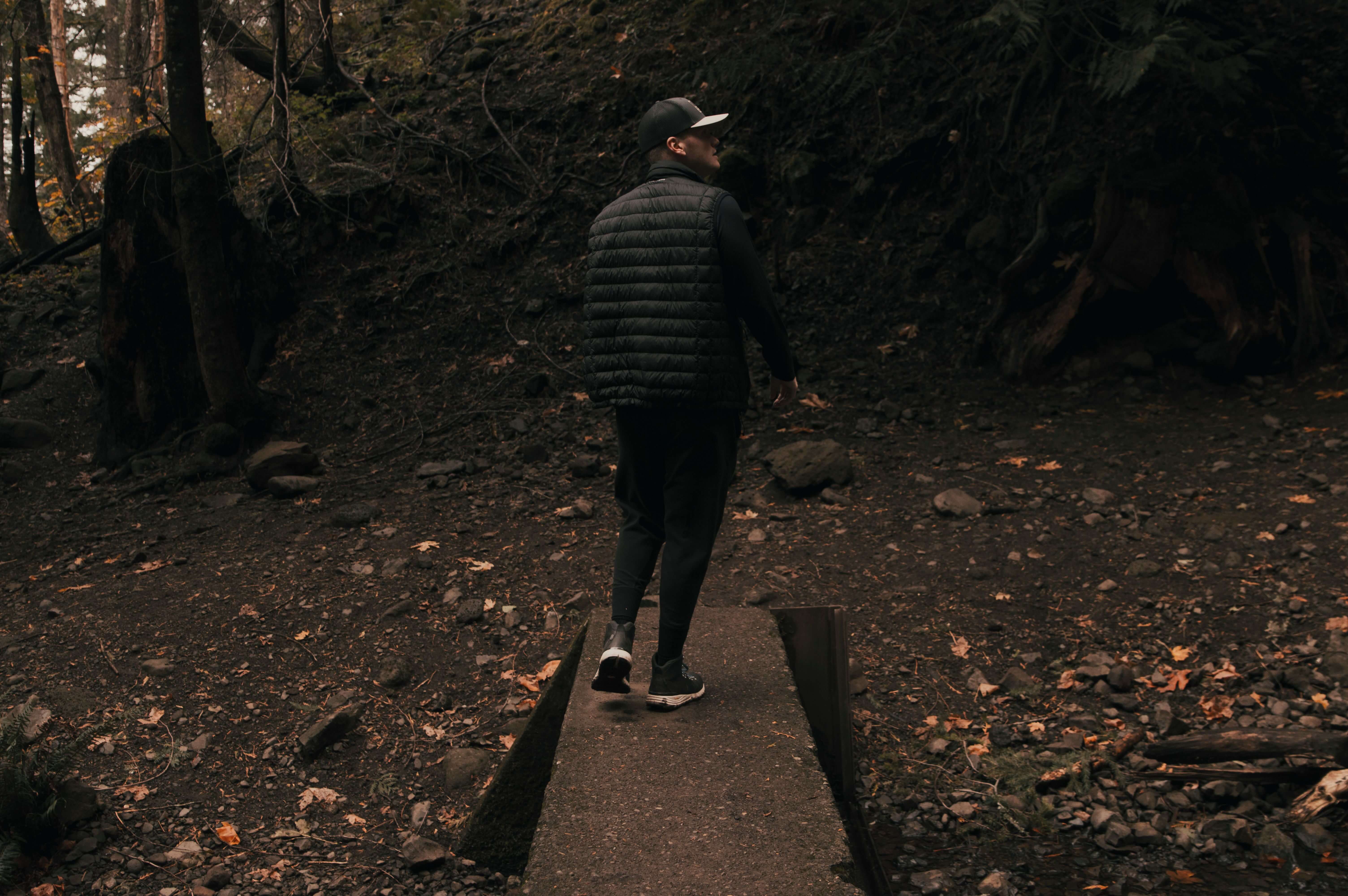 Man in a black puffer vest and cap walking away from camera along a narrow concrete path through a dark autumn forest | tapping for trauma | therapeutic coaching online | international trauma therapist | Los Angeles | Seattle | Chicago