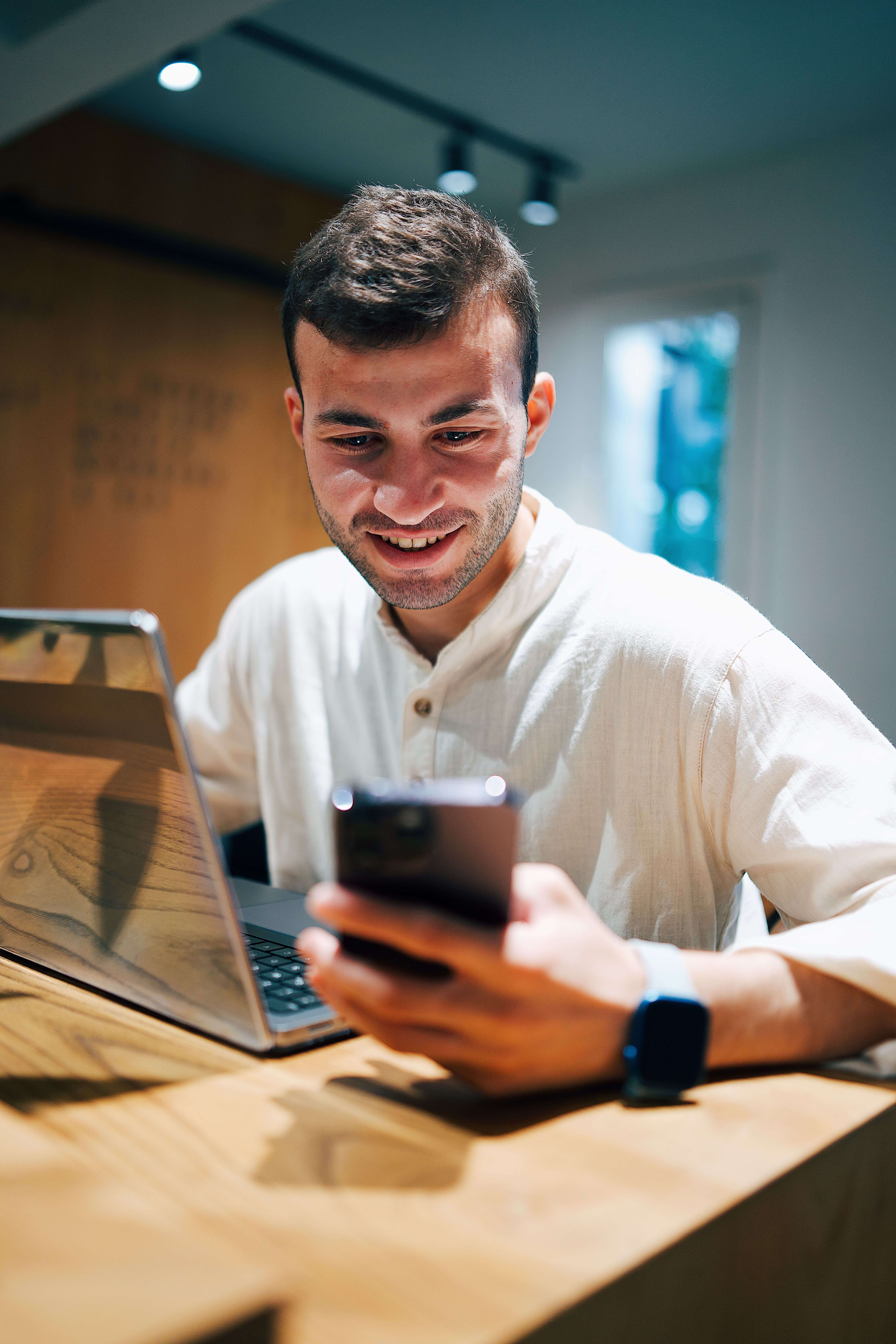 Young man in a white linen shirt smiling at his phone while sitting at a wooden desk with a laptop open beside him | executive mental health | therapy for entrepreneurs | EMDR therapy online | United States | Portugal | Italy