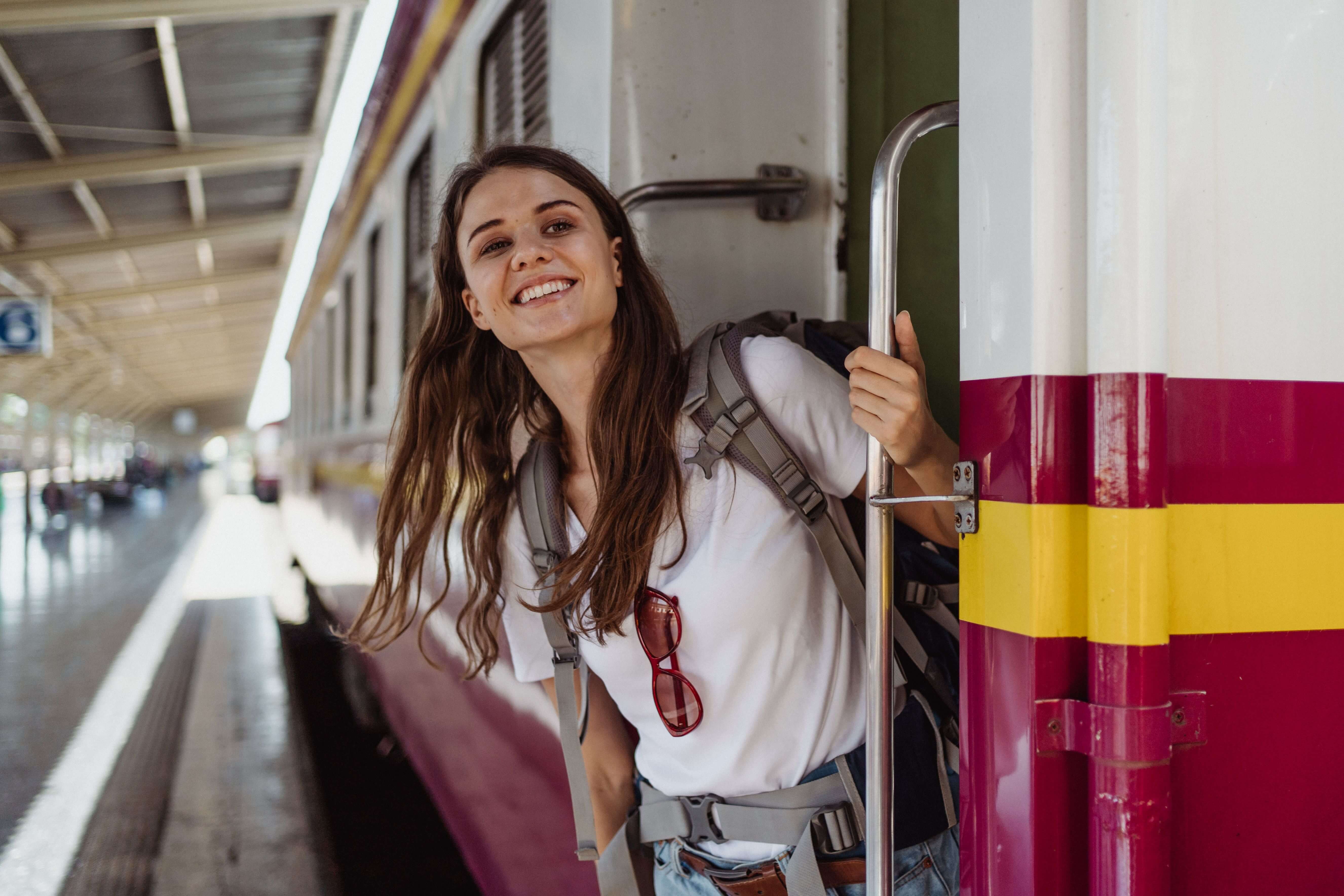 Smiling woman with a backpack leaning out of a train door at a station platform in bright daylight | therapy for expats | expat friendships | international therapist | Portugal | Italy | United States