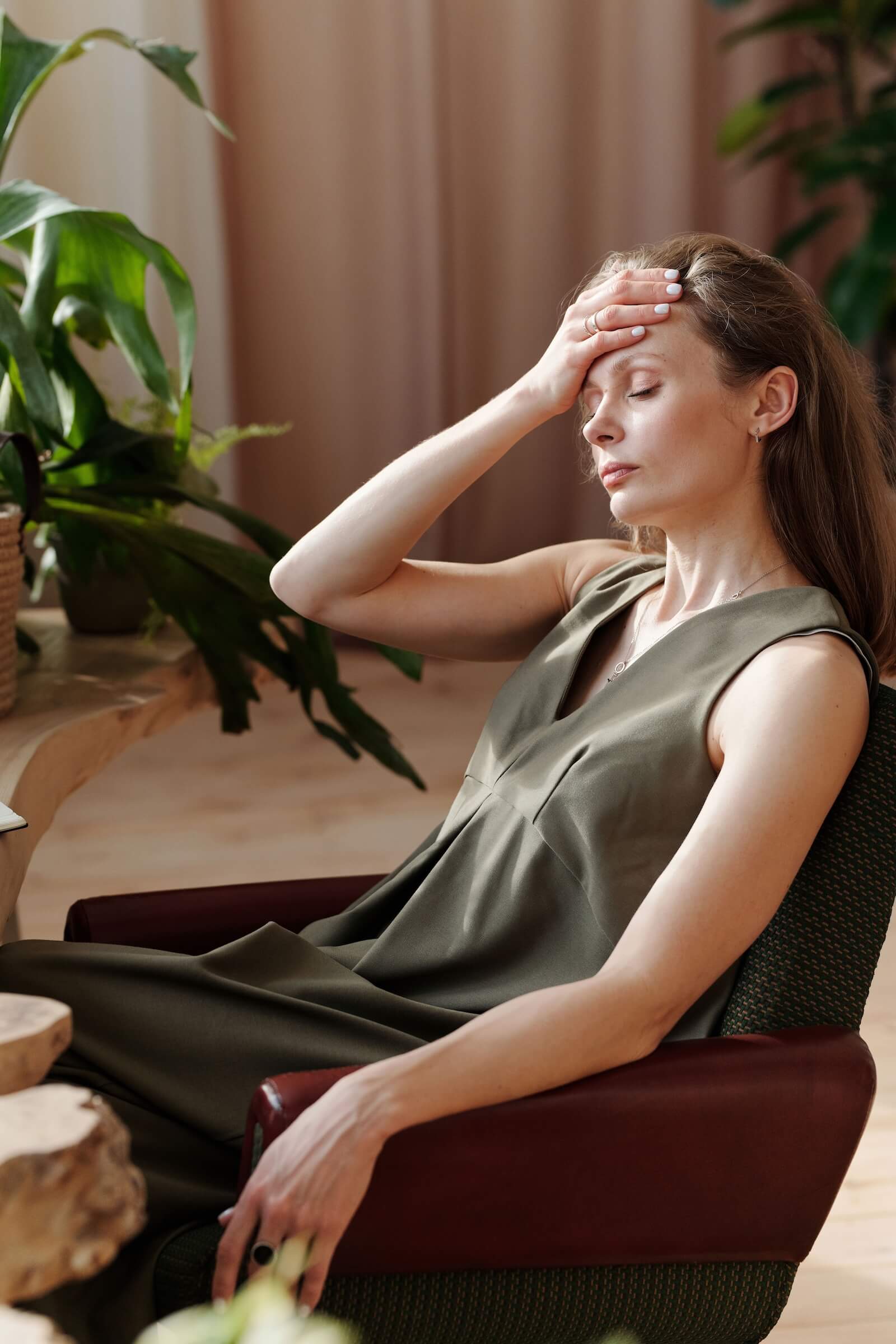 Woman in an olive green dress sitting in a chair with eyes closed and hand pressed to her forehead in a warm sunlit room with plants | tapping therapy | therapeutic coaching | international trauma therapist | Seattle | New York City | Chicago